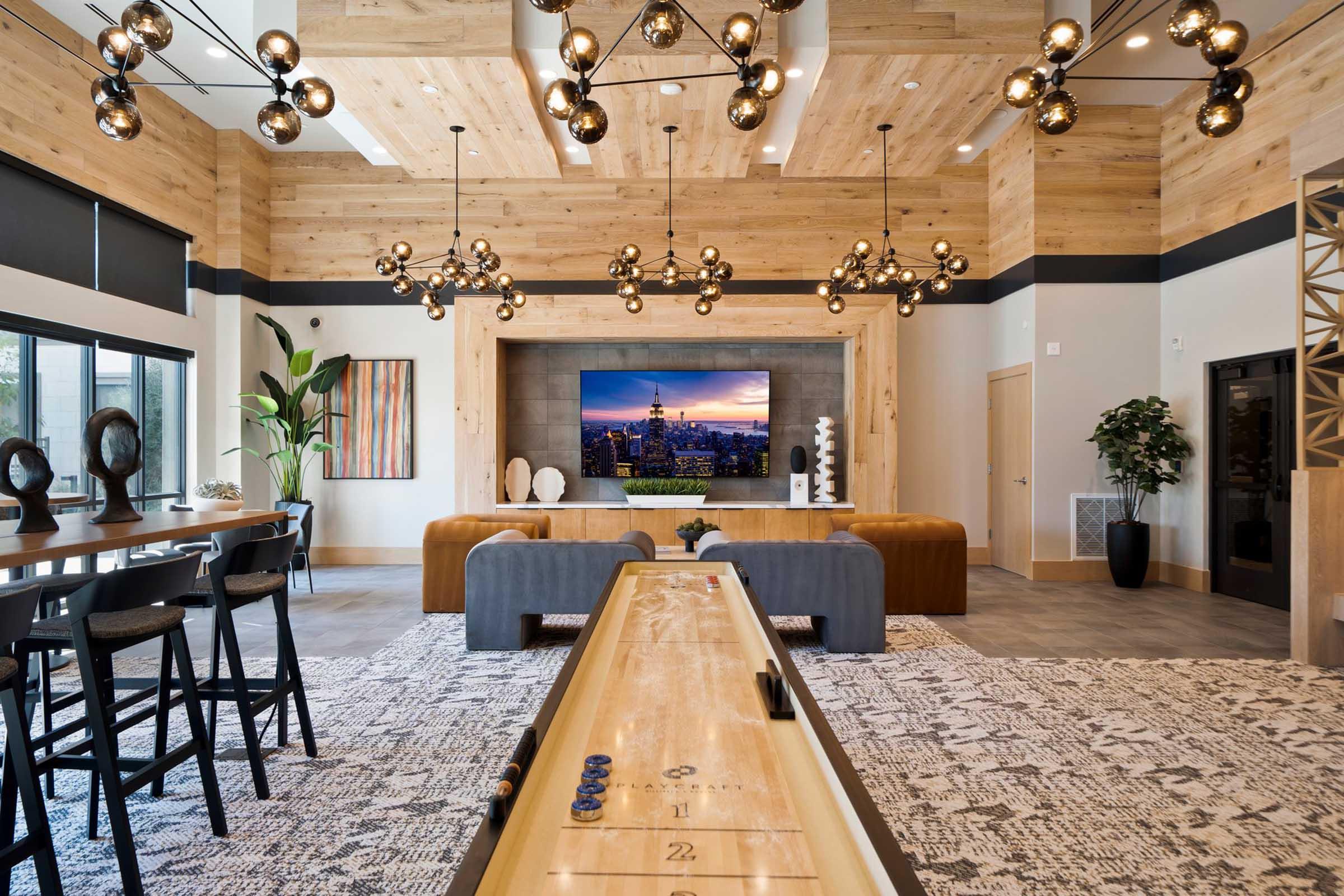 A modern lounge area featuring wooden accents, stylish lighting fixtures, and a large screen displaying a cityscape. There is a shuffleboard table in the center, surrounded by comfortable seating. The decor includes plants and artwork, creating an inviting atmosphere for socializing or relaxation.