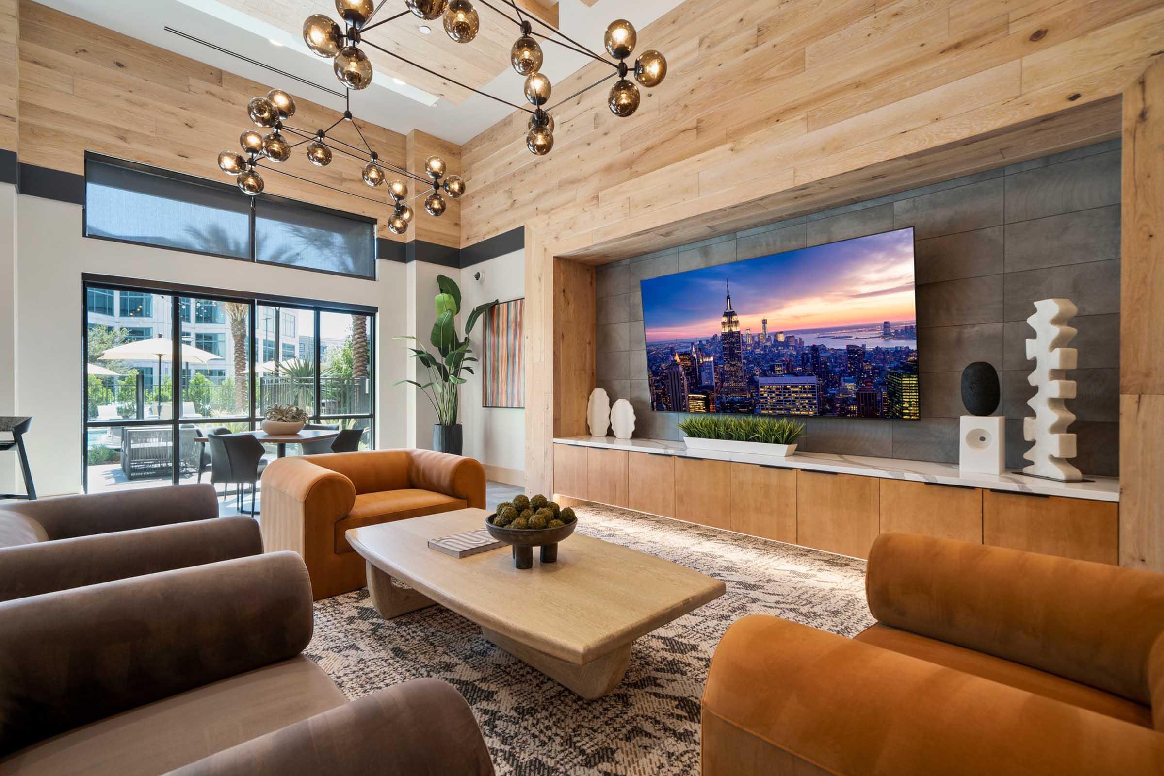 Modern living room interior featuring a wooden accent wall, a large flat-screen TV displaying a city skyline, stylish seating with brown and gray armchairs, a wooden coffee table with decorative items, and large windows that provide natural light and views of an outdoor space.