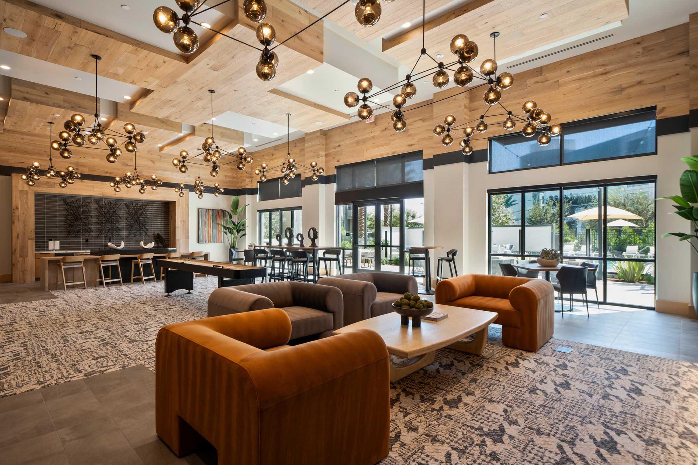 A modern interior space featuring a spacious lounge area with stylish furniture, including a brown sofa and coffee table. The ceiling is adorned with unique light fixtures, and large windows provide natural light. An outdoor patio with umbrellas and greenery is visible through the glass doors.