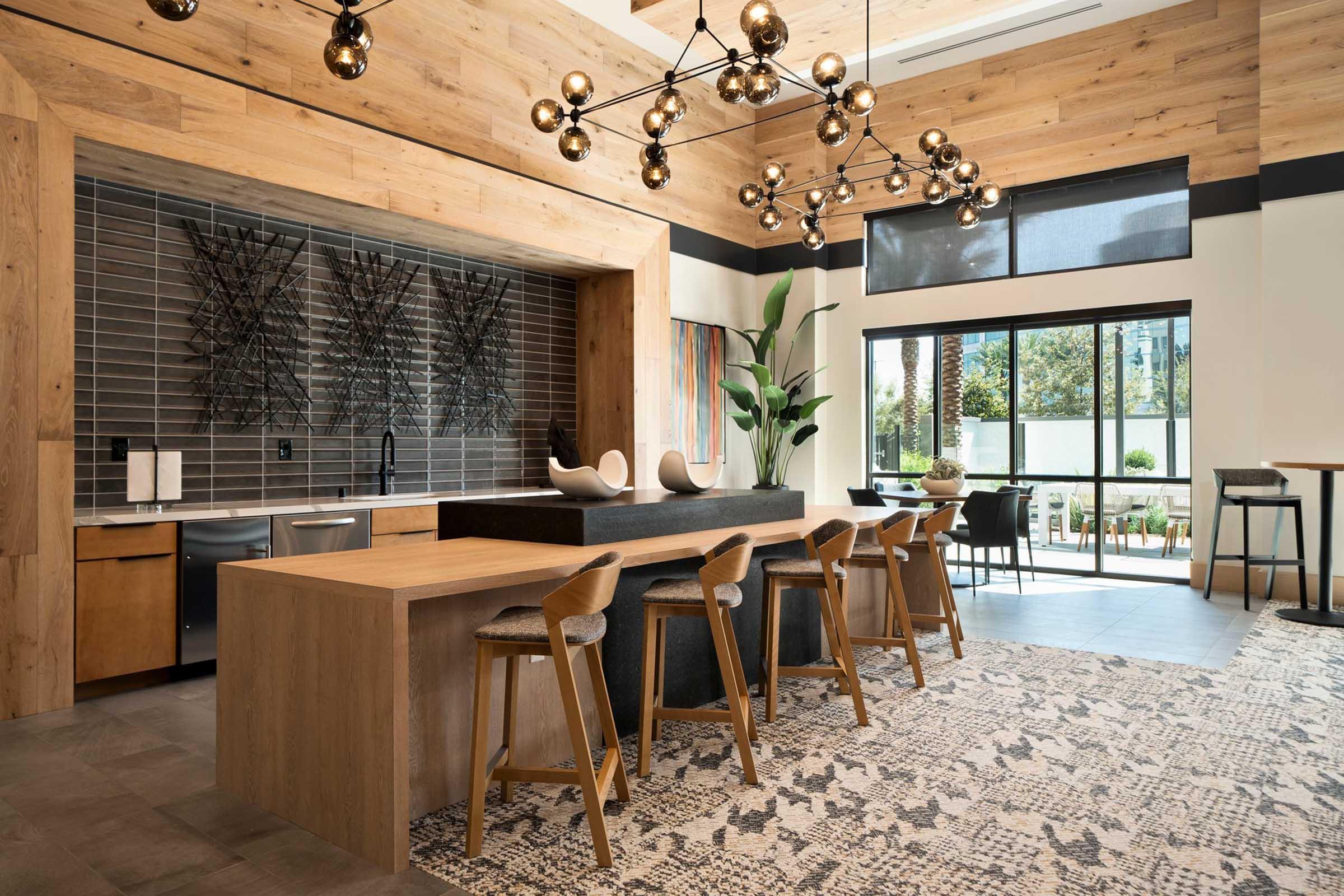 A modern interior of a stylish kitchen and dining area featuring wooden accents, a large island with bar stools, geometric lighting fixtures, and decorative plants. Large windows allow natural light, and the décor includes textured wall art and a cozy rug.