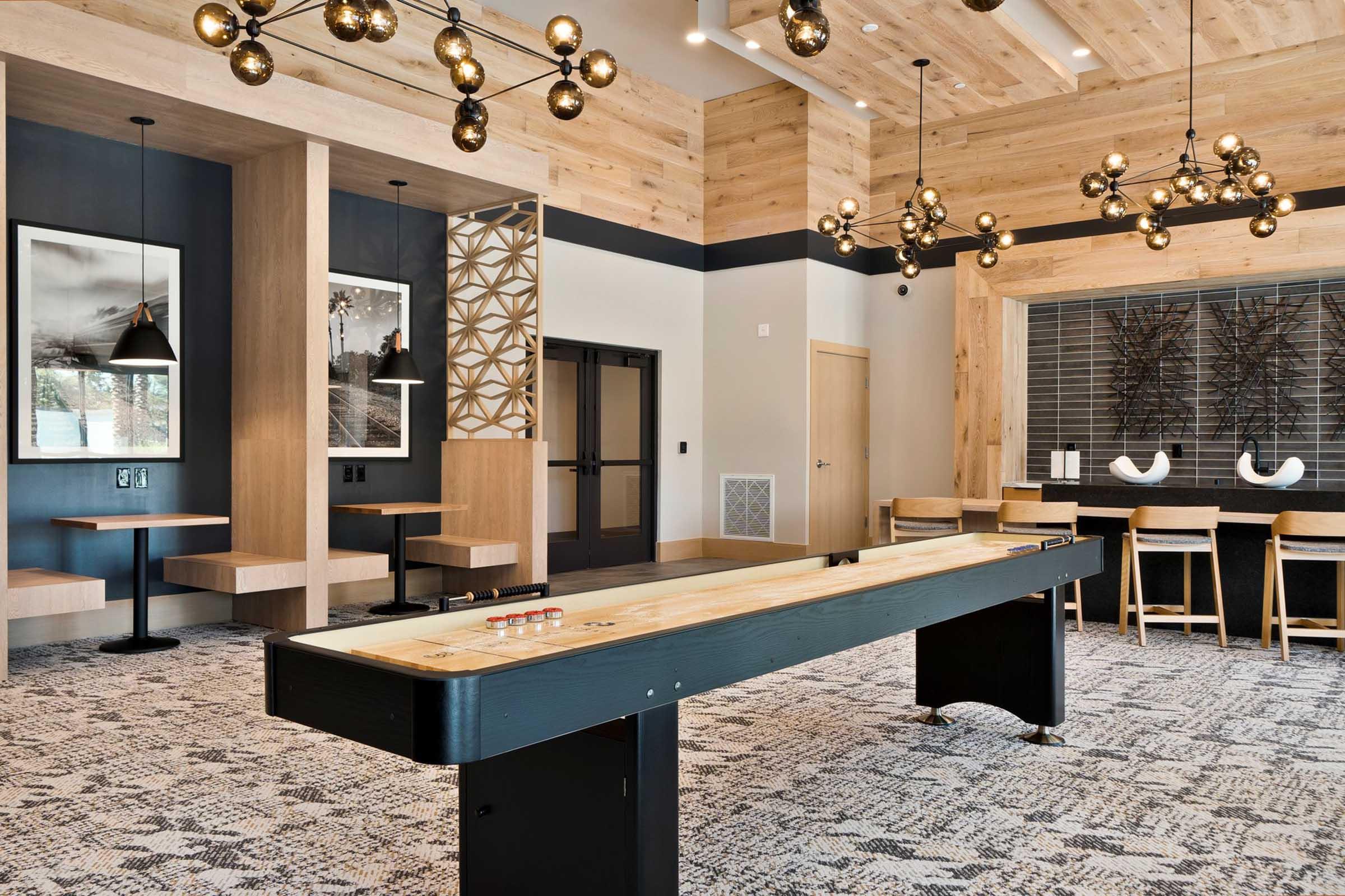 A modern lounge area featuring a shuffleboard table in the center, surrounded by stylish wooden decor. Soft pendant lighting hangs from the ceiling, while wall-mounted art and seating areas create a welcoming atmosphere. The flooring has a patterned carpet, adding to the contemporary design.