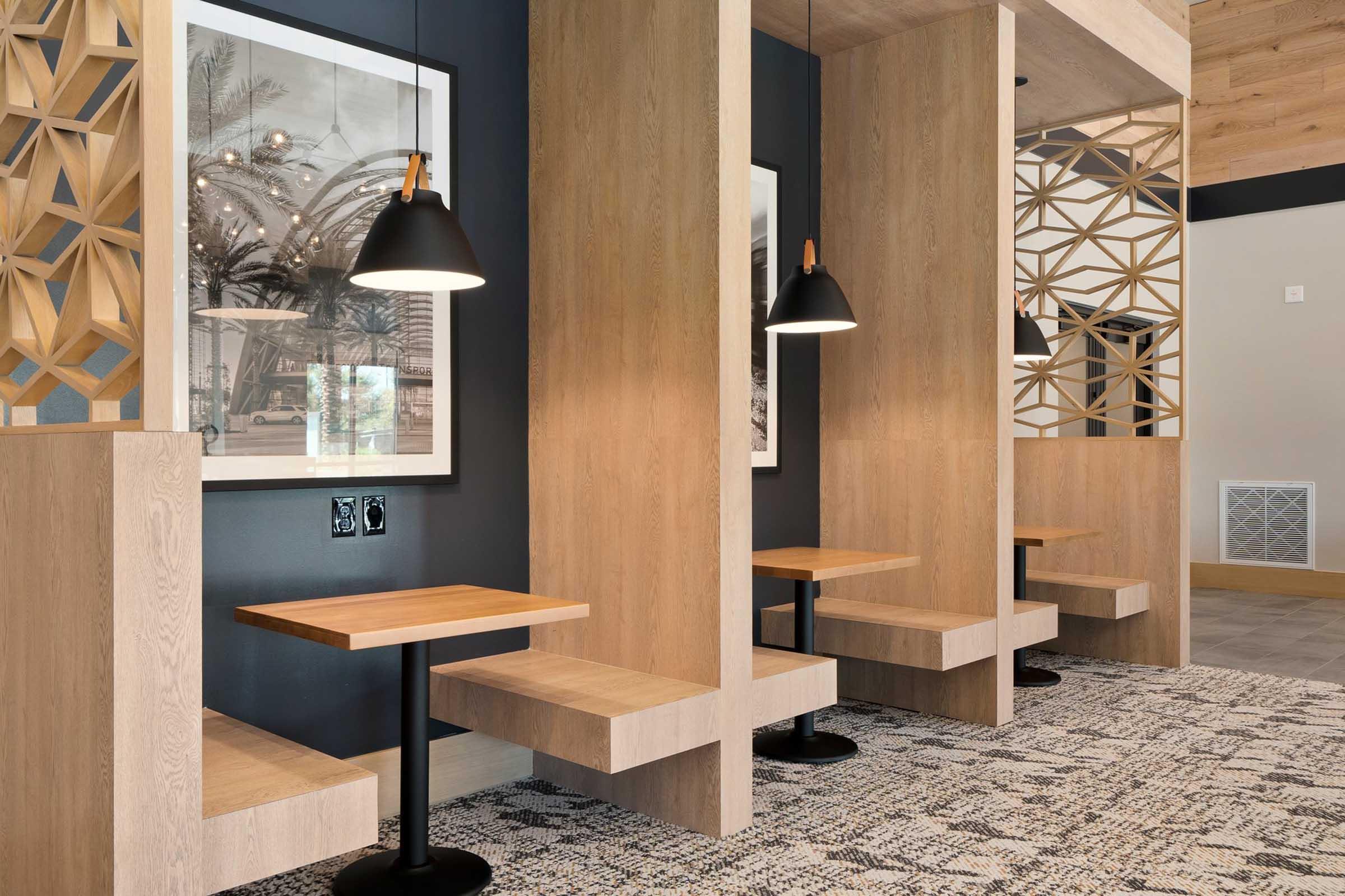 A modern, stylish interior featuring wooden booths with low tables. The space showcases decorative wall panels and pendant lighting, creating a warm ambiance. The flooring is textured, and there are framed photographs on the walls, adding visual interest to the contemporary design.