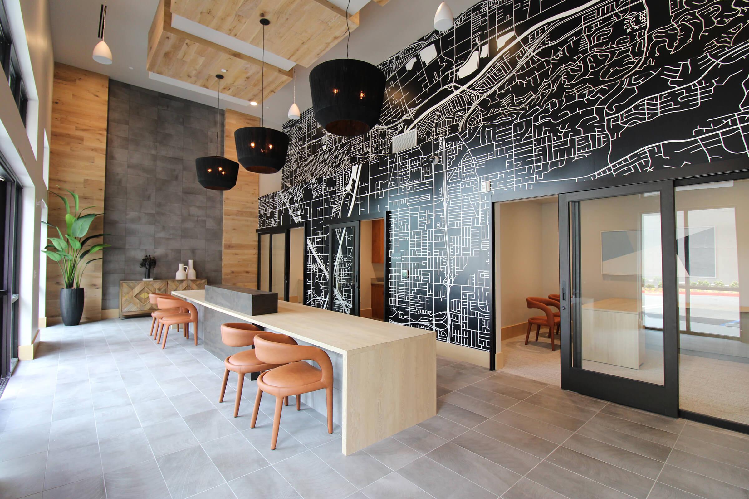 Modern interior of a lobby featuring a large wooden table with brown chairs, stylish pendant lights, and a striking black and white map mural on the wall. The space includes large windows for natural light and a decorative plant in a planter, creating an inviting and contemporary atmosphere.