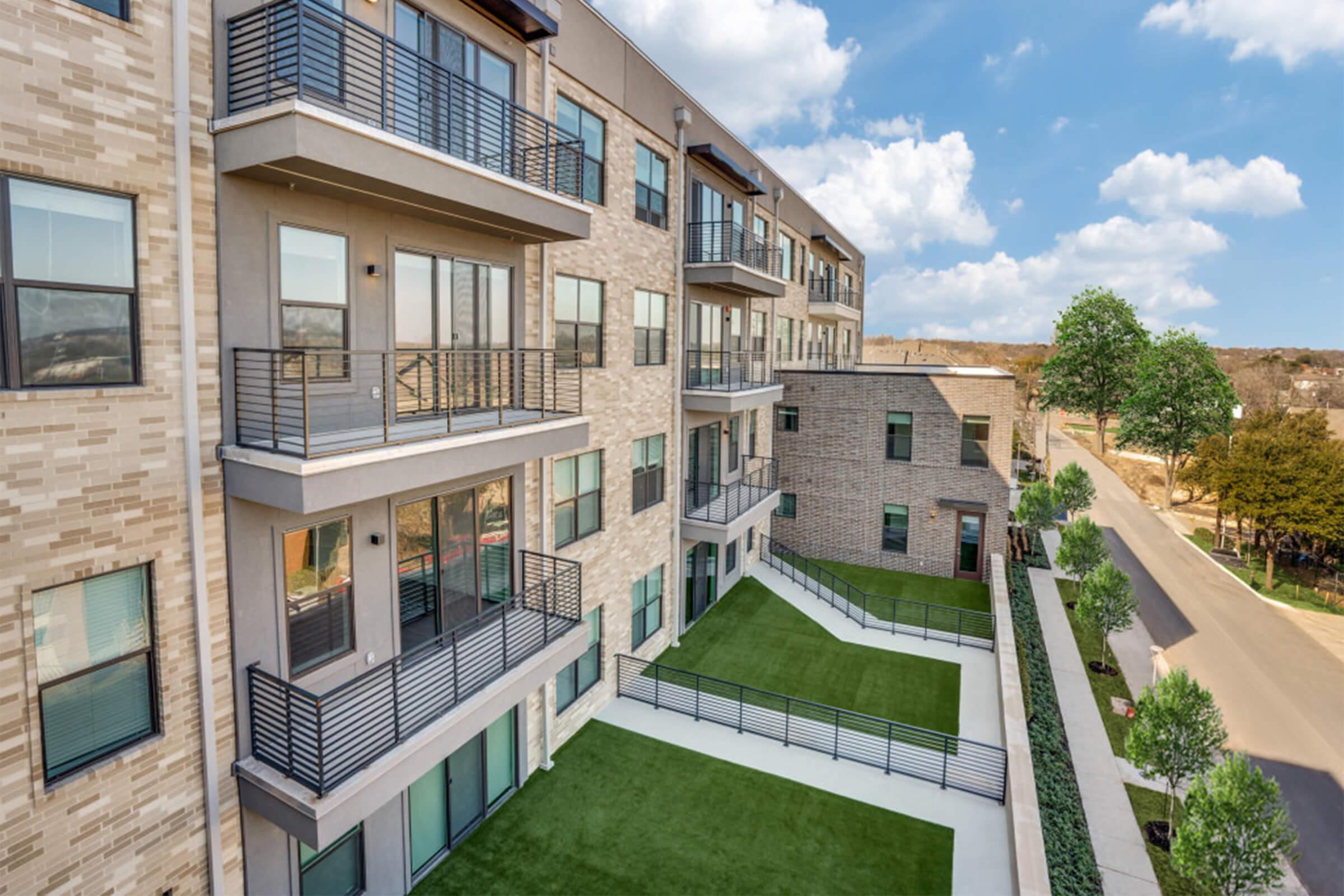 A modern apartment building featuring multiple balconies, large windows, and a green outdoor space. The architecture includes a mix of brick and contemporary design elements. Surrounding the building are neatly landscaped areas with trees and a pathway along the side.