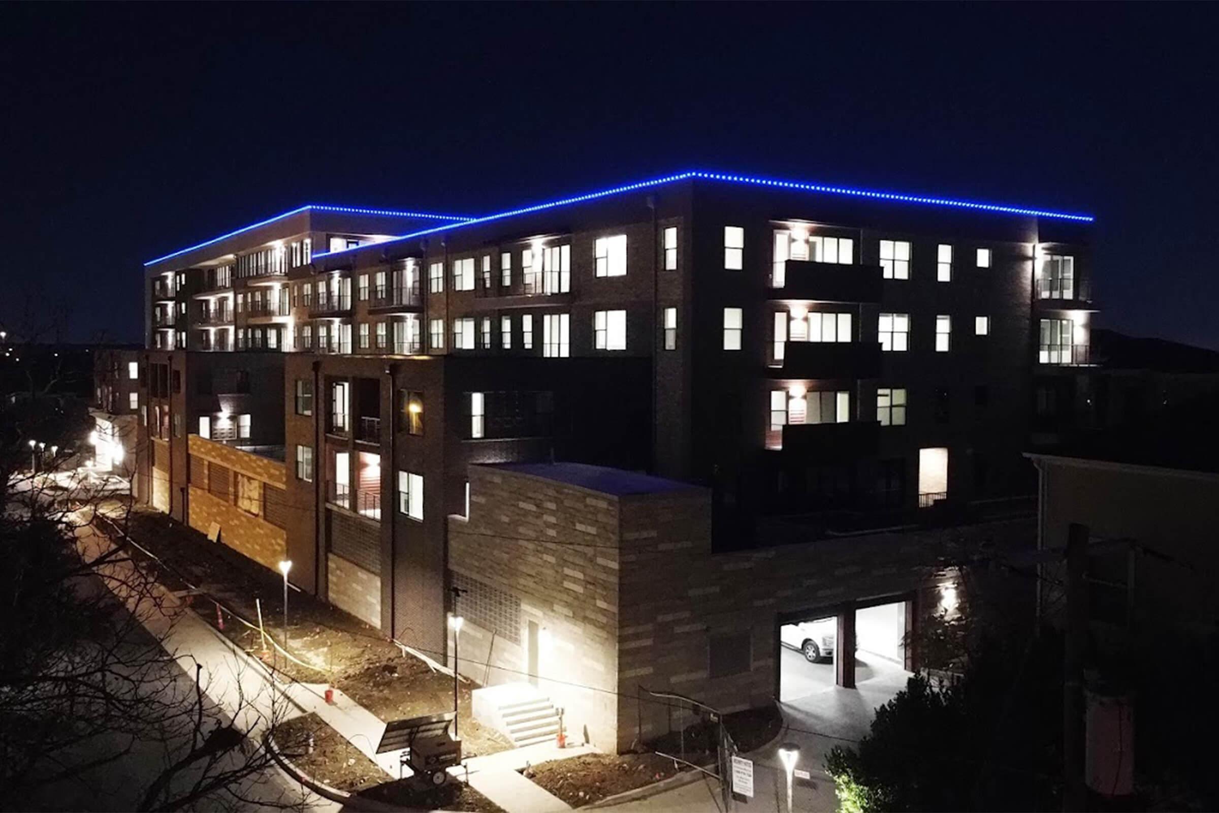 A modern apartment building at night, illuminated with blue LED lights along the roofline. The structure features multiple stories with windows brightly lit. Surrounding pathways are softly lit, creating a welcoming atmosphere in the evening.