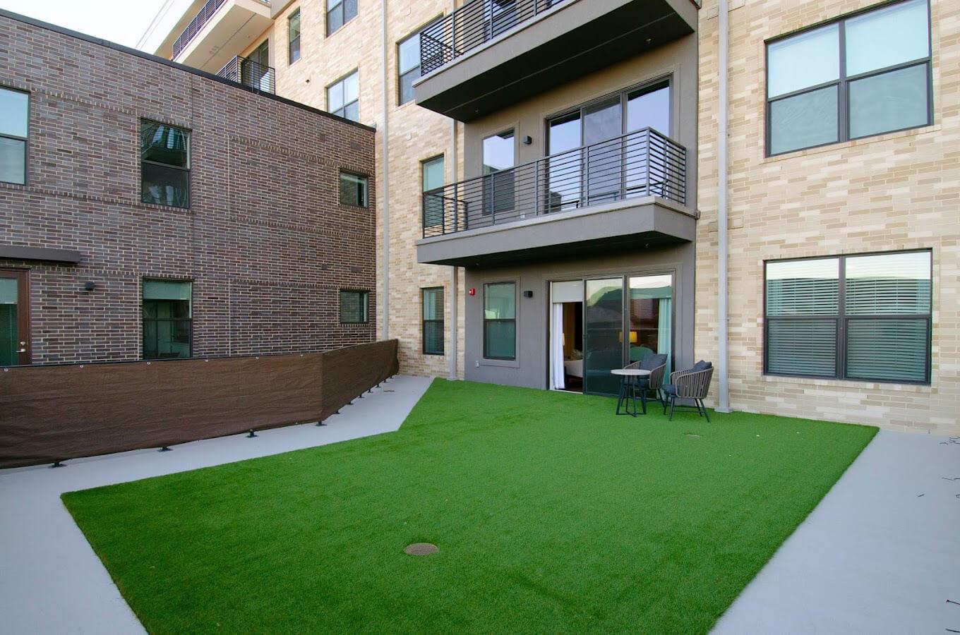 A modern apartment balcony with artificial grass, featuring a small seating area with two chairs and a table. The balcony looks out onto a courtyard with neighboring buildings visible in the background. The architecture includes a mix of brick and light-colored siding.