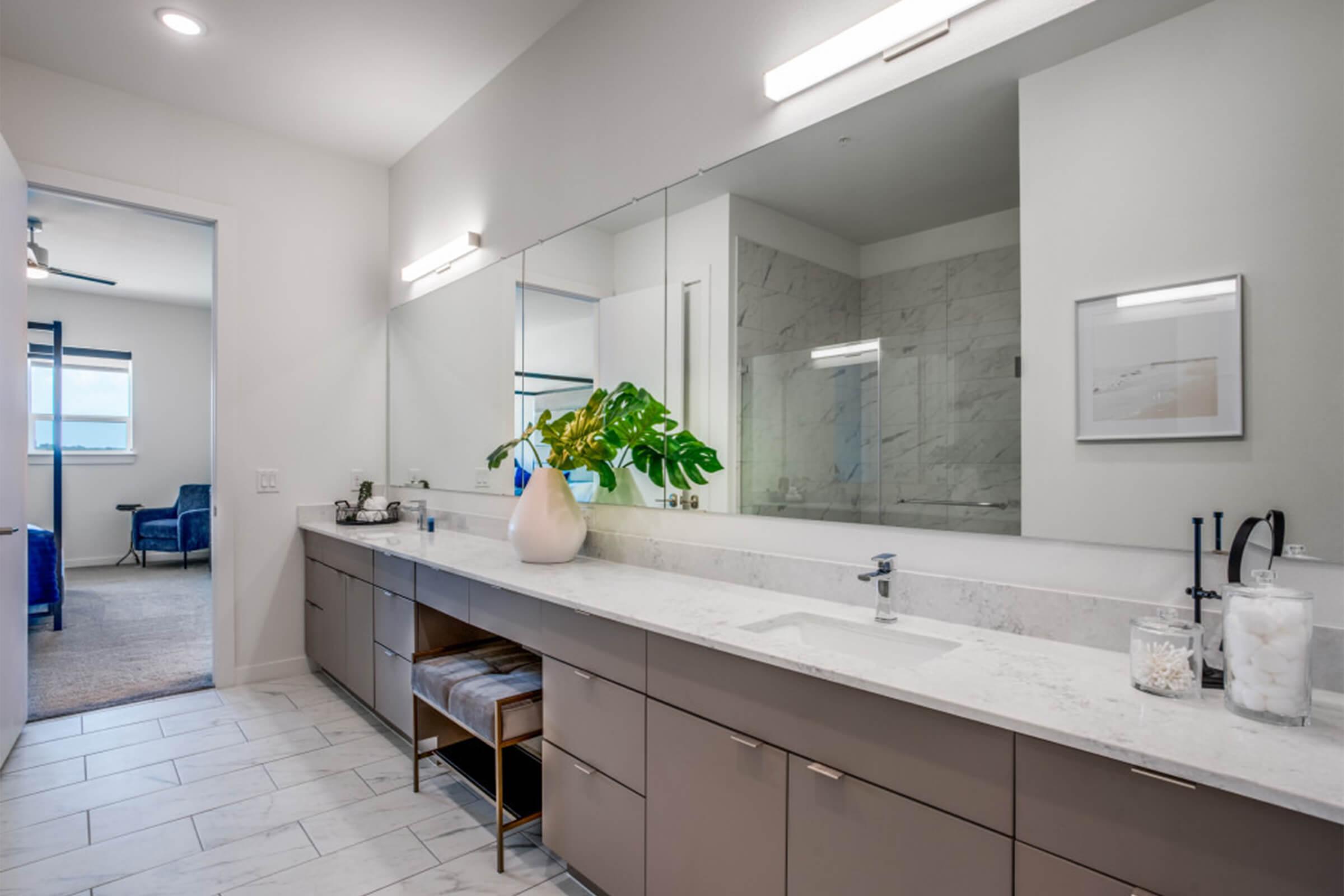 Modern bathroom featuring a large vanity with dual sinks, a spacious mirror, and stylish lighting. A neutral color palette is complemented by a decorative plant. The design includes sleek cabinetry and a glass shower visible in the background, with a door leading to another room.