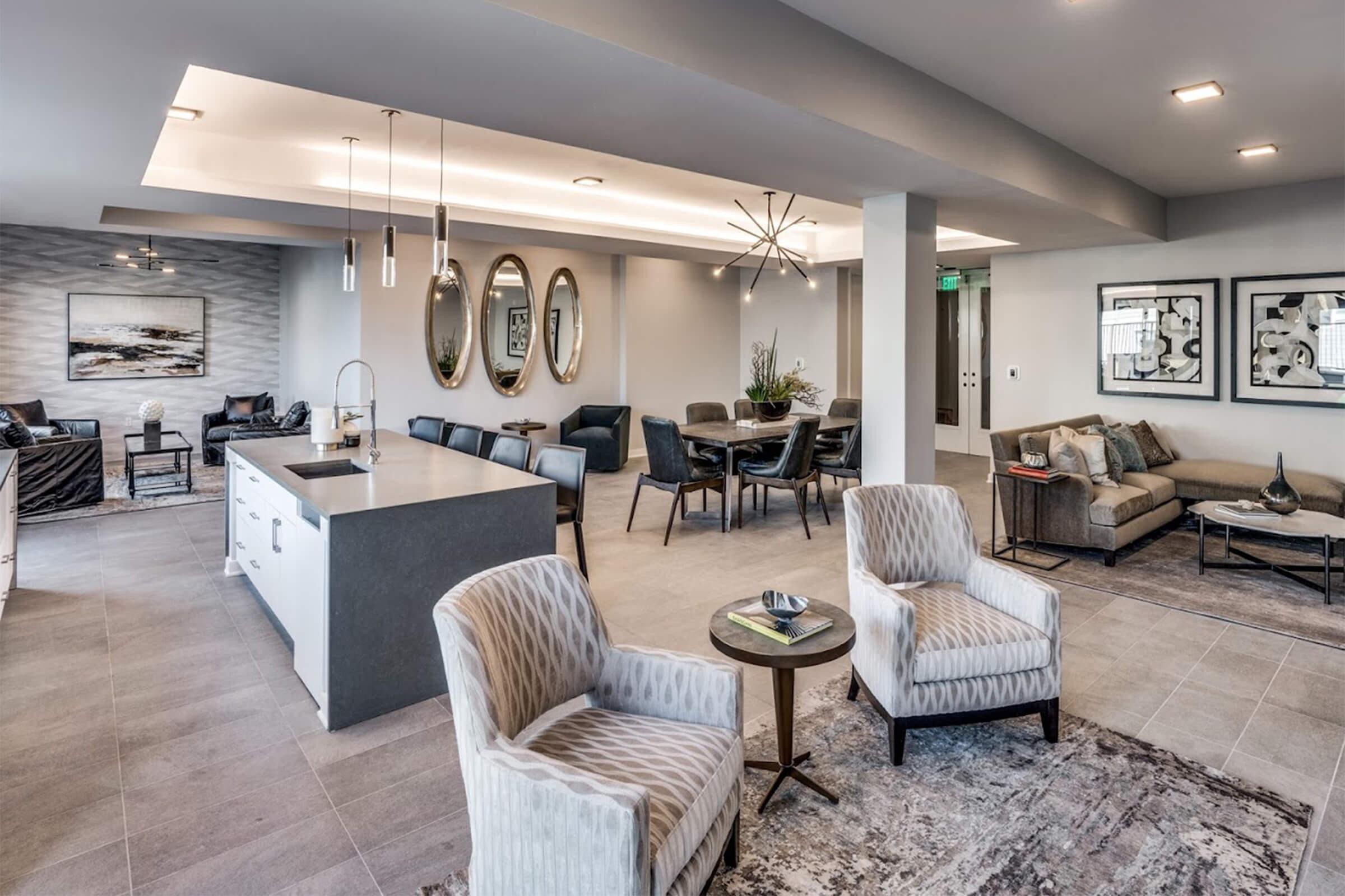 Modern apartment interior showcasing a spacious common area with stylish furniture. Features include two plush armchairs, a sleek kitchen island, dining tables, and contemporary decor. Large mirrors and artwork adorn the walls, creating a bright and inviting atmosphere.