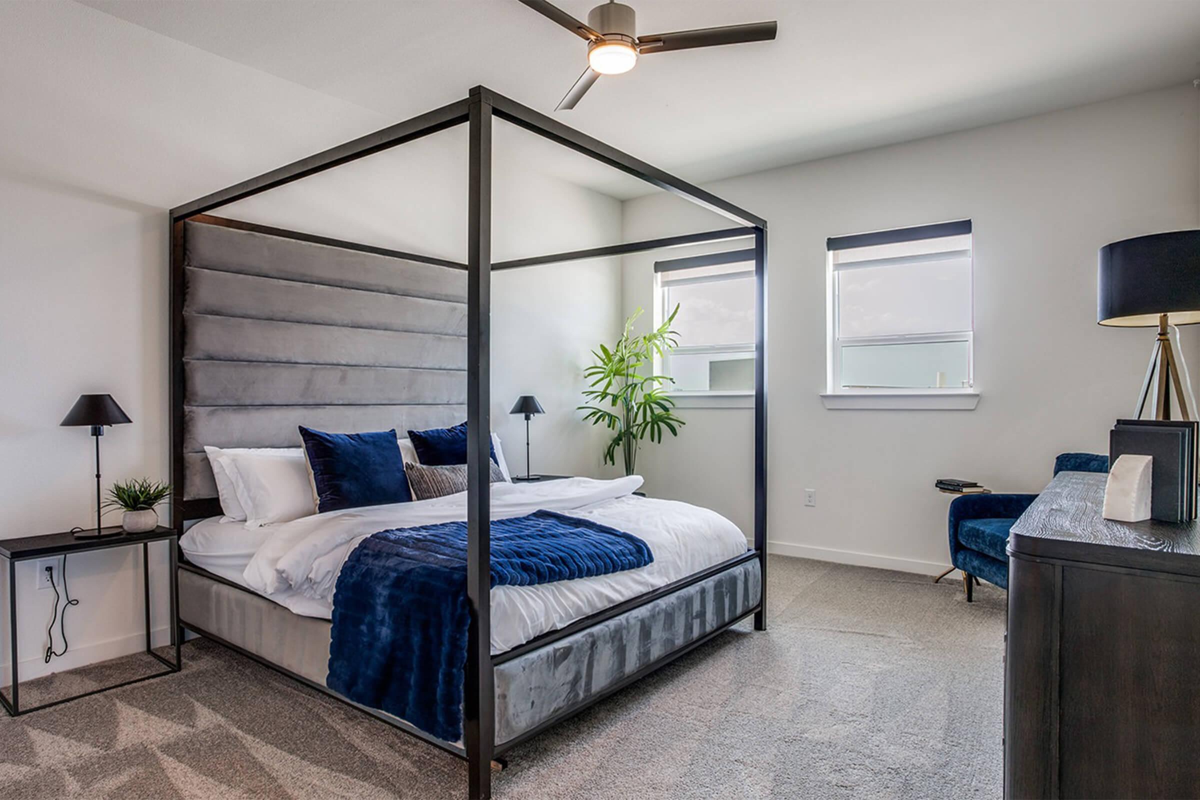 A modern bedroom featuring a four-poster bed with a gray upholstered headboard, crisp white bedding, and decorative blue pillows. The room includes a blue accent chair, a lamp, and a green plant by the window. Natural light fills the space, creating a serene atmosphere on a beige carpet.