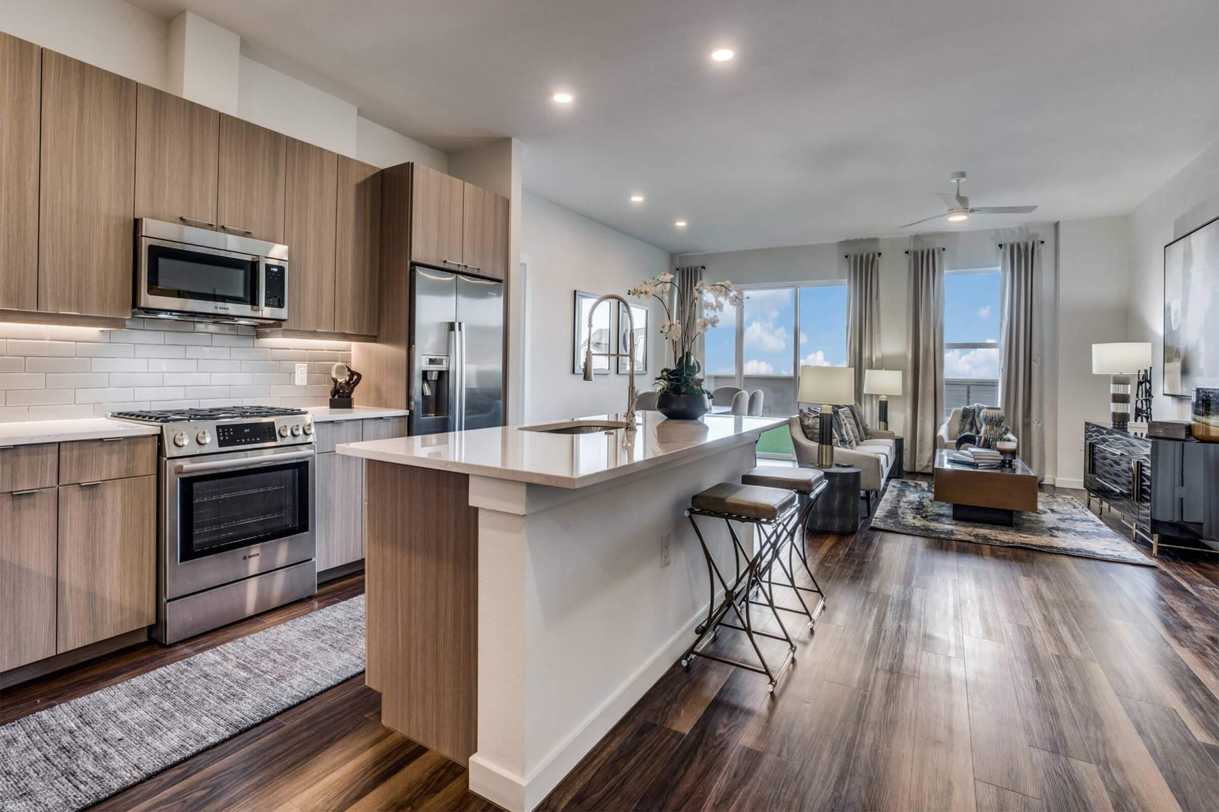 Modern kitchen and living area in an apartment. The kitchen features sleek wooden cabinets, stainless steel appliances, and a large island with bar stools. The living room has a comfortable sofa, coffee table, and large windows with natural light, complemented by stylish decor and a neutral color palette.