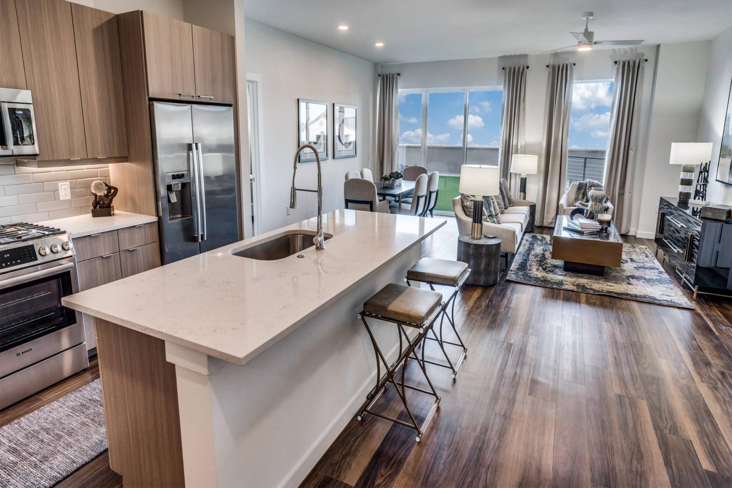 Modern open-concept apartment interior featuring a stylish kitchen with a central island, stainless steel appliances, and wood cabinetry. The living area has comfortable seating, a coffee table, and large windows that let in natural light, with a dining table visible in the background.