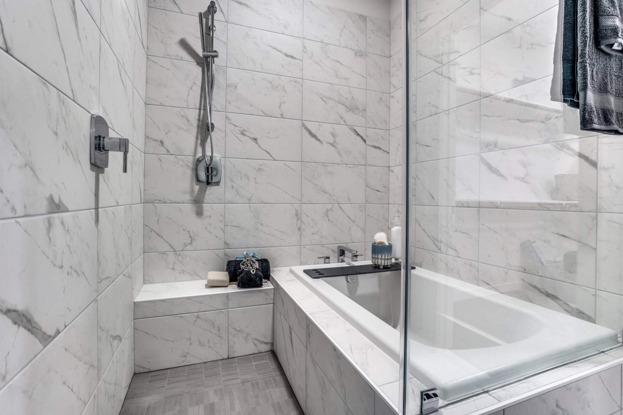 Modern bathroom featuring a white marble tile design with a glass shower enclosure. A sleek bathtub is positioned against one wall, with soap and toiletries neatly arranged nearby. Towels hang on a rail, creating a clean and stylish look in the space.