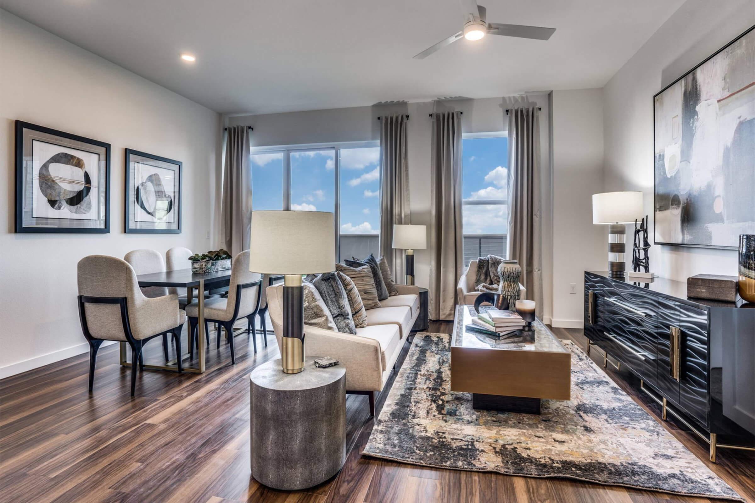 A modern living room featuring a comfortable sofa, stylish armchairs, and a chic coffee table. The space is decorated with framed artwork, a floor lamp, and a large window allowing natural light to illuminate the room. Wooden flooring adds warmth, complementing the contemporary design.