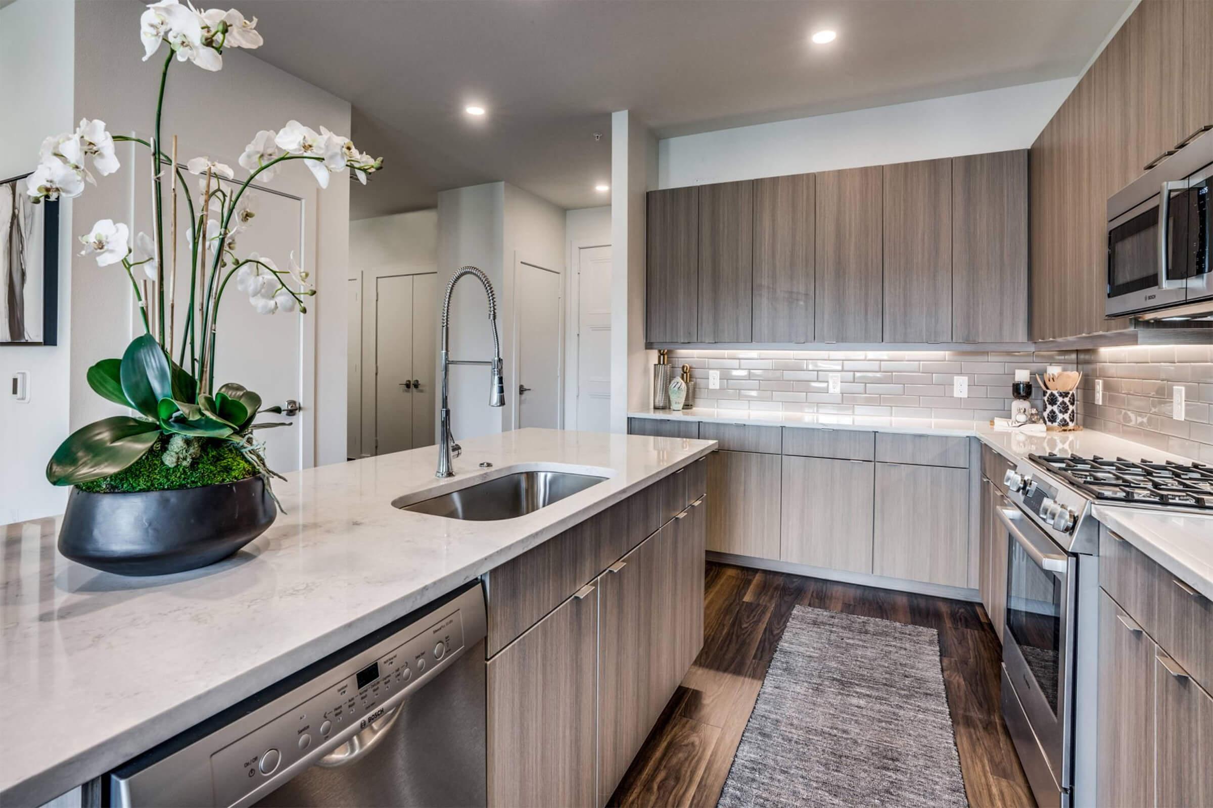 A modern kitchen featuring sleek wooden cabinets, a large island with a stainless steel sink, and a built-in dishwasher. The countertop is made of light-colored stone. A decorative arrangement of white orchids adds a touch of elegance. Soft lighting creates a warm ambiance.