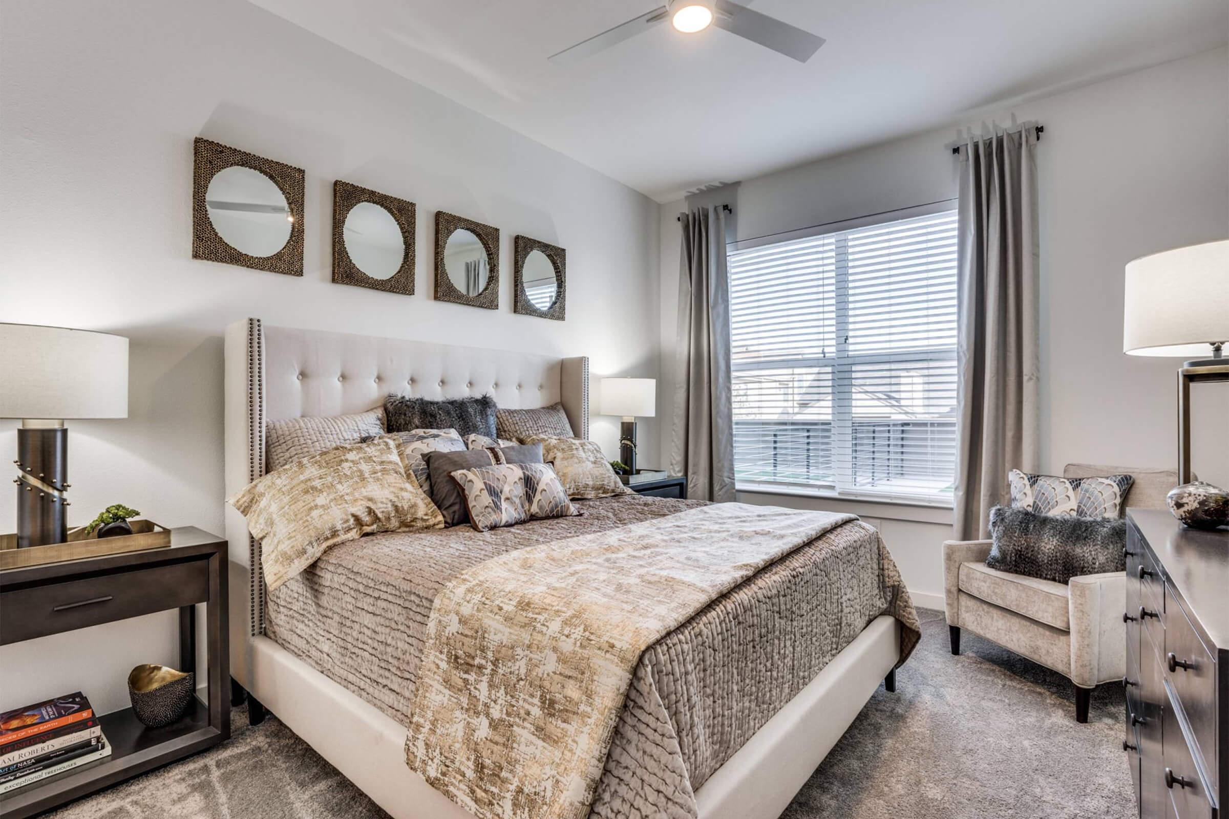 A stylish bedroom featuring a large bed with decorative pillows, complemented by a tufted headboard. Three round mirrors hang above the bed, and a window with sheer curtains lets in natural light. A bedside table with a lamp and a cozy armchair complete the inviting atmosphere.