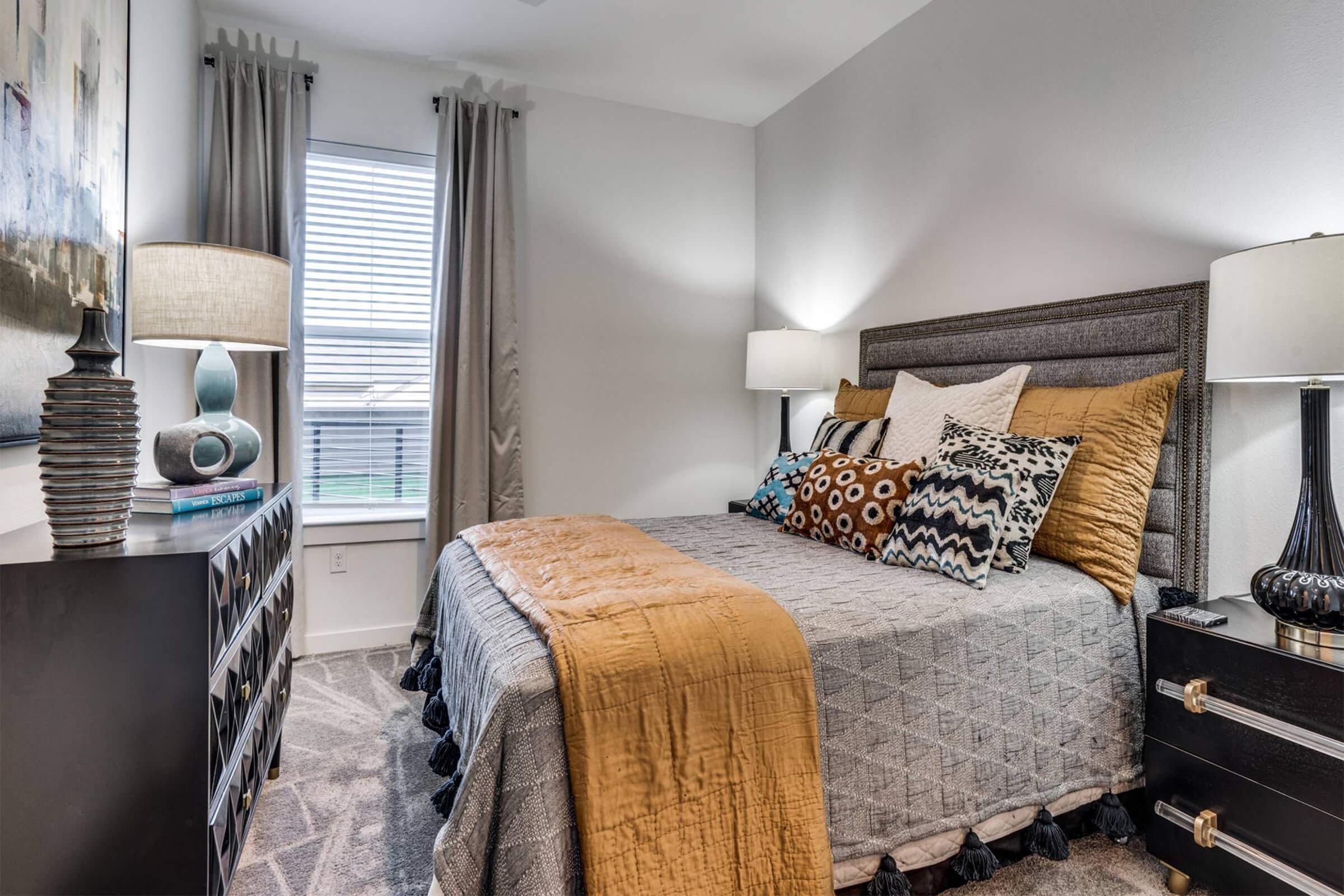 A cozy bedroom featuring a neatly made bed with decorative pillows, a warm-colored throw, and stylish bedding. The room has two lamps on bedside tables, large windows with sheer curtains, and a modern dresser on one side, creating a welcoming and comfortable atmosphere.