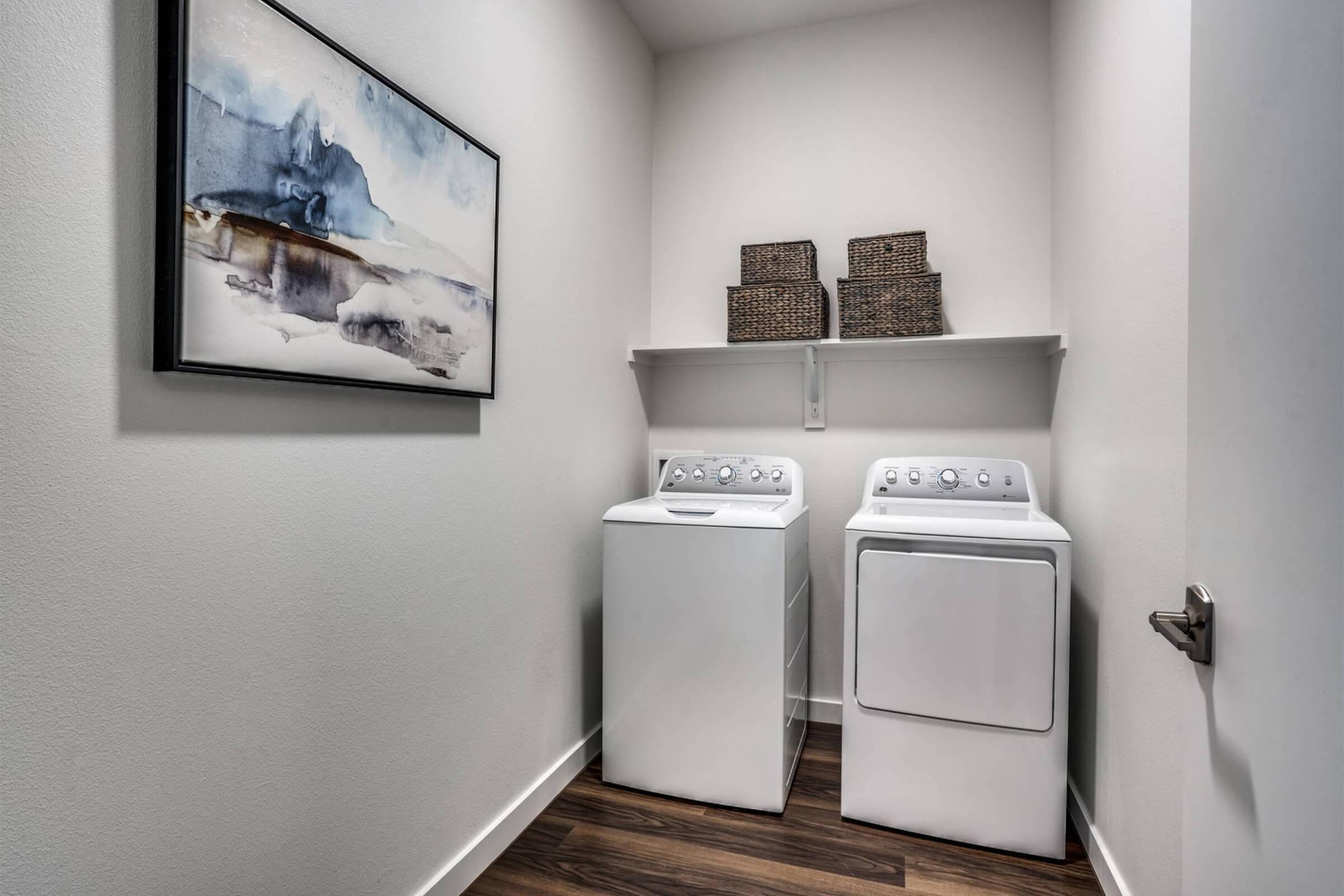 A clean laundry room featuring a stacked washer and dryer against a light-colored wall. Above the appliances, there is a shelf with decorative woven baskets. A framed abstract painting hangs on the wall, adding a touch of modern decor to the space. The flooring is dark wood, complementing the overall design.