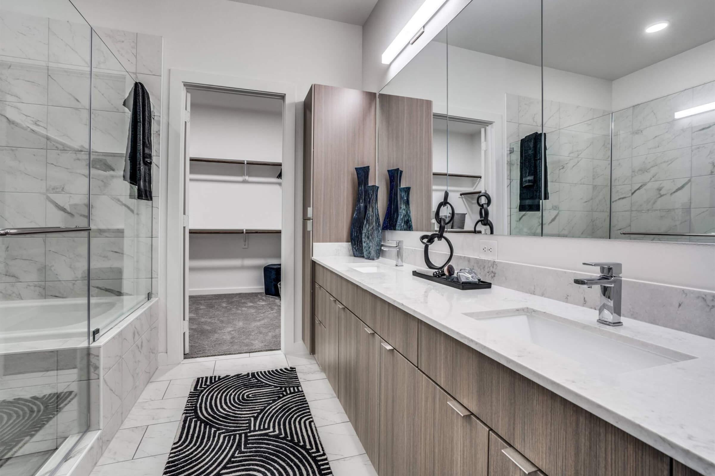 A modern bathroom featuring a double sink vanity with wood cabinetry, a large mirror, and elegant decor. There is a glass-enclosed shower, a bathtub, and a stylish black and white patterned rug. A walk-in closet is visible in the background, and the space has bright lighting and a clean, contemporary design.