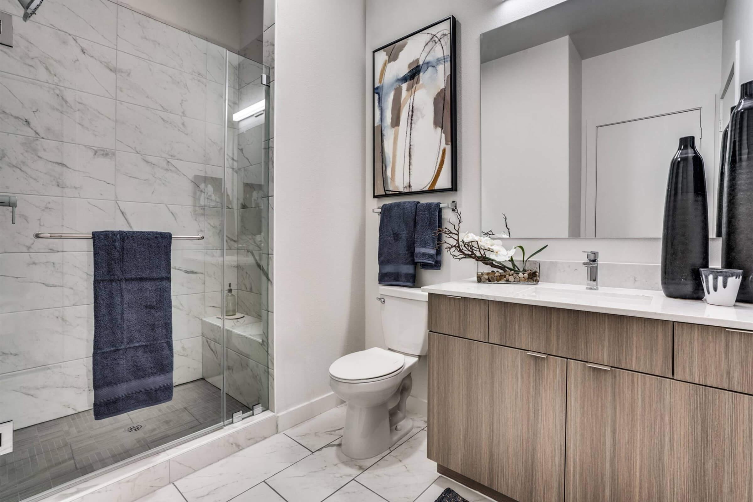 Modern bathroom featuring a glass shower with marble walls, a wooden vanity with a white countertop, a toilet, and decorative elements including towels, a vase with flowers, and artwork on the wall. The overall design is sleek and contemporary, with neutral colors and clean lines.