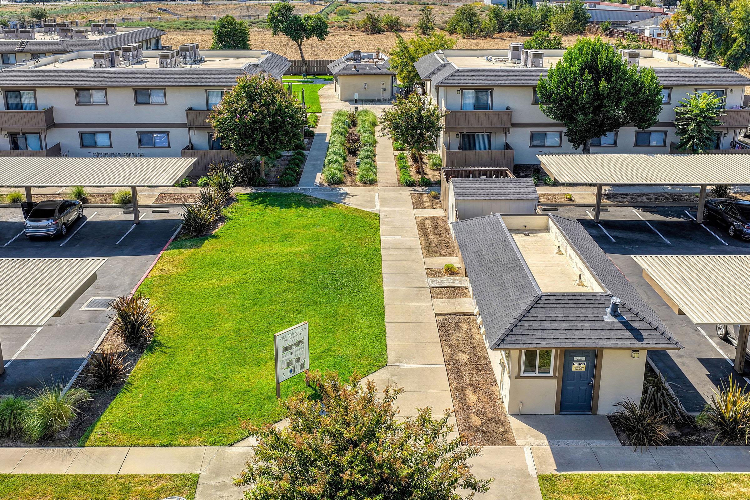 Aerial view of a residential complex featuring two-story buildings with parking spaces under shaded structures. A well-maintained lawn and landscape separate the buildings, with pathways leading through the greenery. The area is surrounded by open fields and trees in the background.