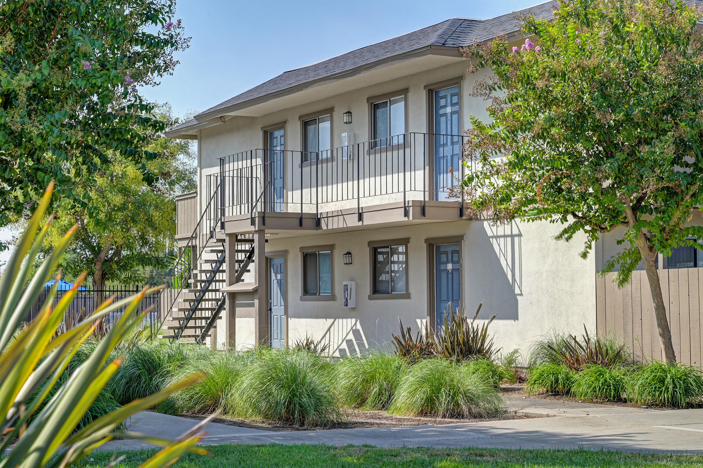 Two-story building with a set of stairs leading to the upper level. The facade features several windows with railings. Surrounding the building are well-maintained grassy areas and bushes. The scene is bright and sunny, showcasing a clear blue sky in the background.