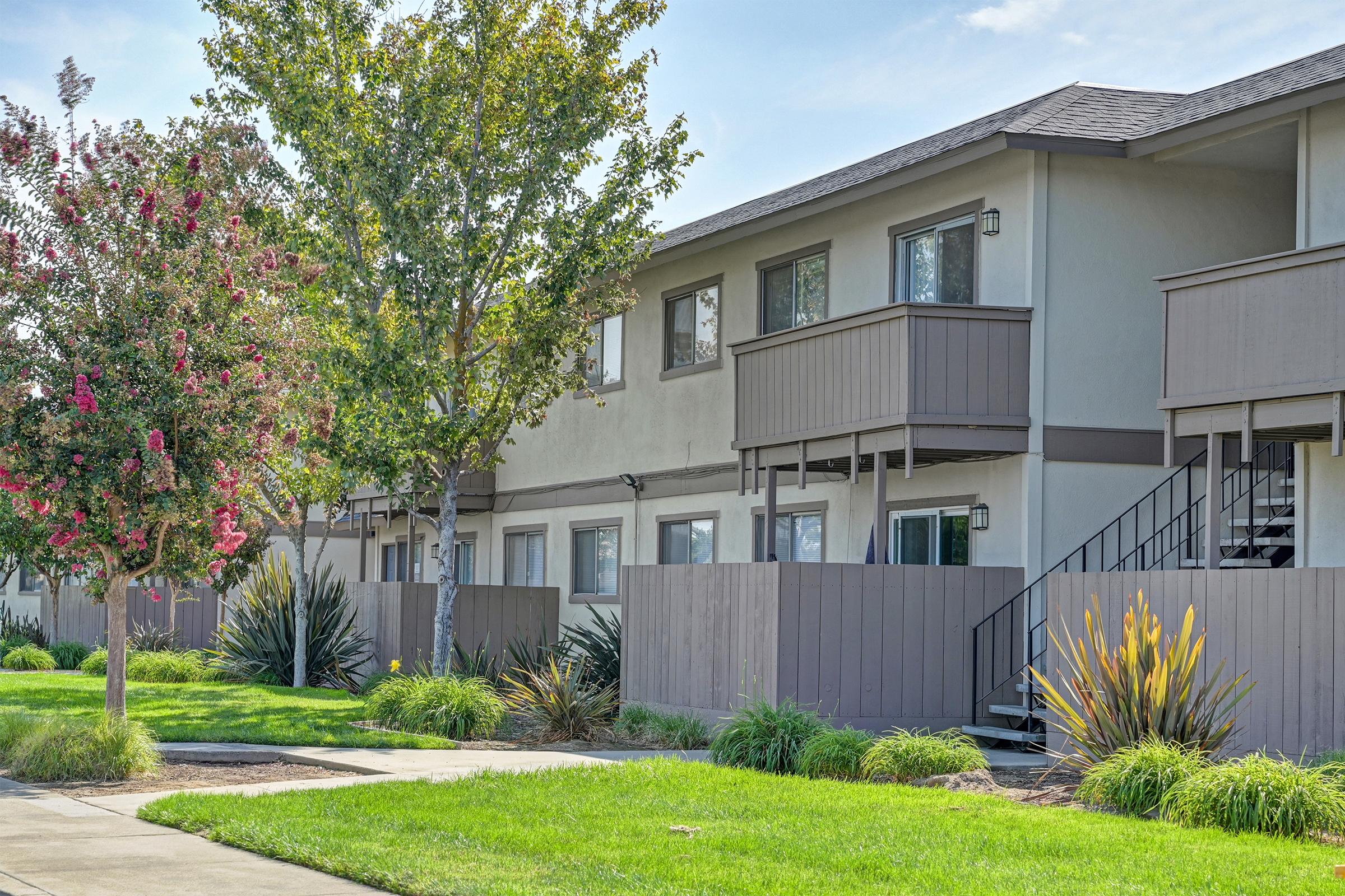 A residential building with multiple units featuring balconies. Lush green grass and colorful shrubs are in the foreground, creating a landscaped area. Trees provide shade along the sidewalk, enhancing the pleasant neighborhood ambiance. The architecture is modern and well-maintained.