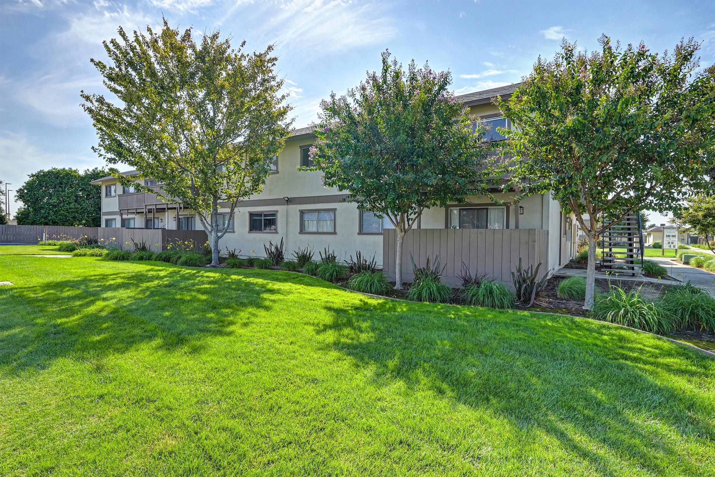 A green lawn in front of a residential building with two stories, surrounded by trees and shrubs. The sky is clear with a few clouds, and there are walkways leading to the building. The scene is bright and well-maintained, showcasing a peaceful outdoor environment.