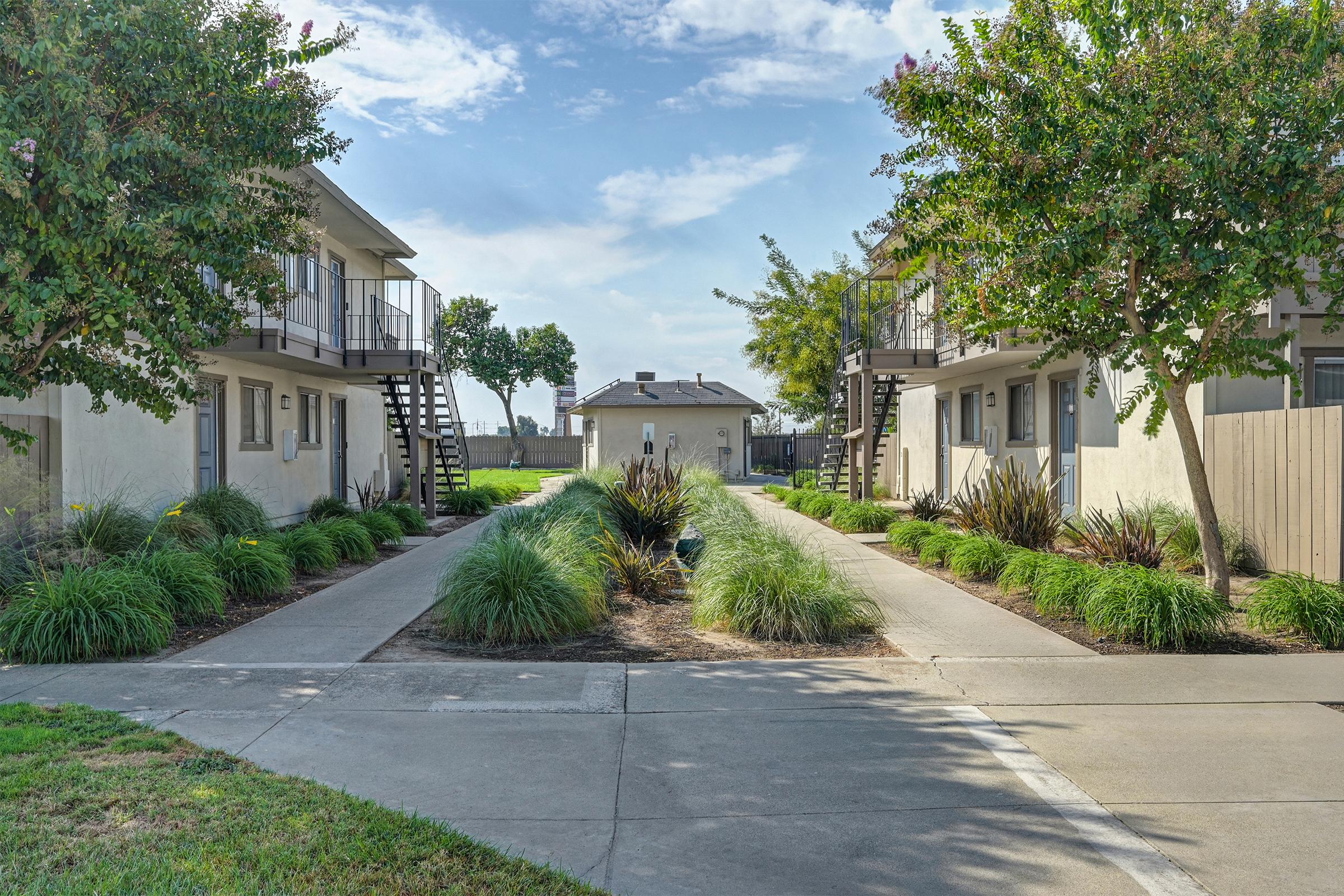 Two sets of two-story apartment buildings flank a well-landscaped pathway. Lush greenery, including ornamental grasses and trees, lines the walkways, which lead to a small, single-story building in the background. The sky is clear with a few clouds, creating a bright and inviting atmosphere.