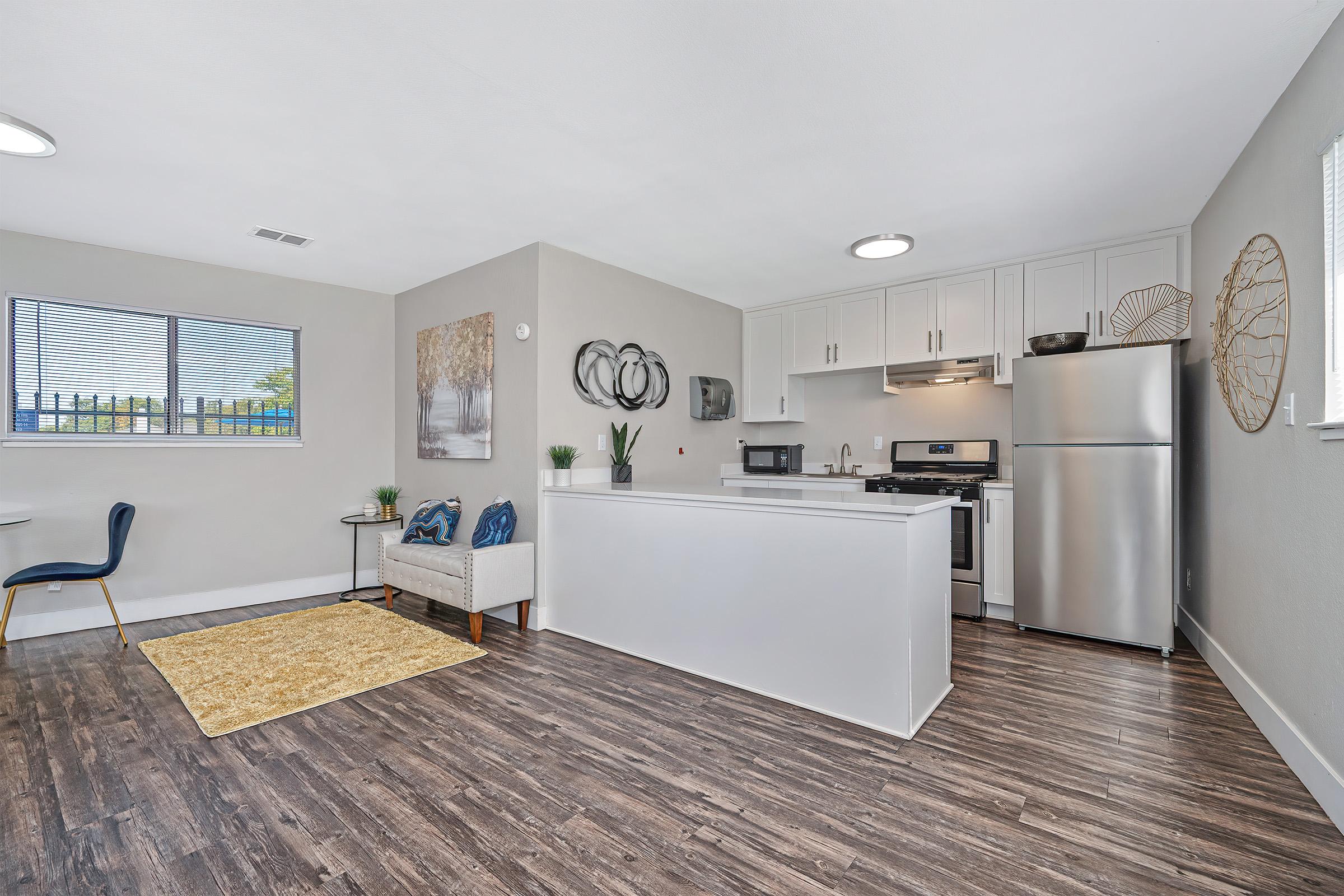 Modern open-concept kitchen and living area featuring stainless steel appliances, a small dining table with a chair, and a cozy seating area. Light-colored walls and wooden flooring enhance the spacious feel. A window lets in natural light, and decorative elements add a touch of style.