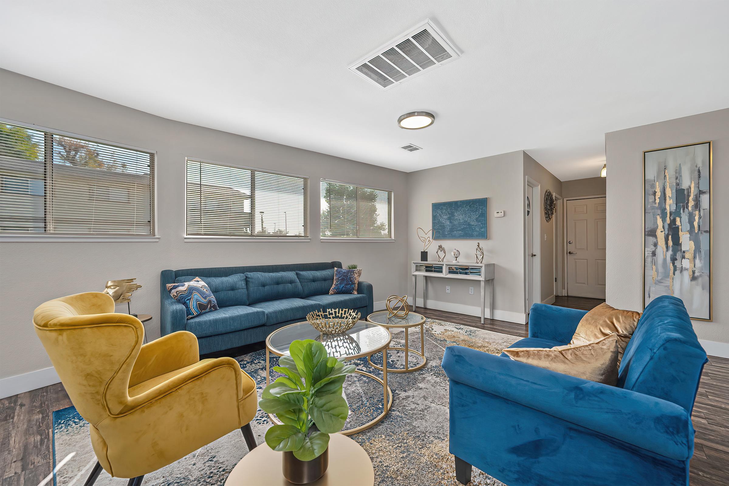 A modern living room featuring a blue sectional sofa, a yellow armchair, and a round coffee table. Large windows allow natural light to brighten the space, highlighting the decorative rug and a small potted plant on the table. A piece of abstract artwork is displayed on the wall, adding a contemporary touch.