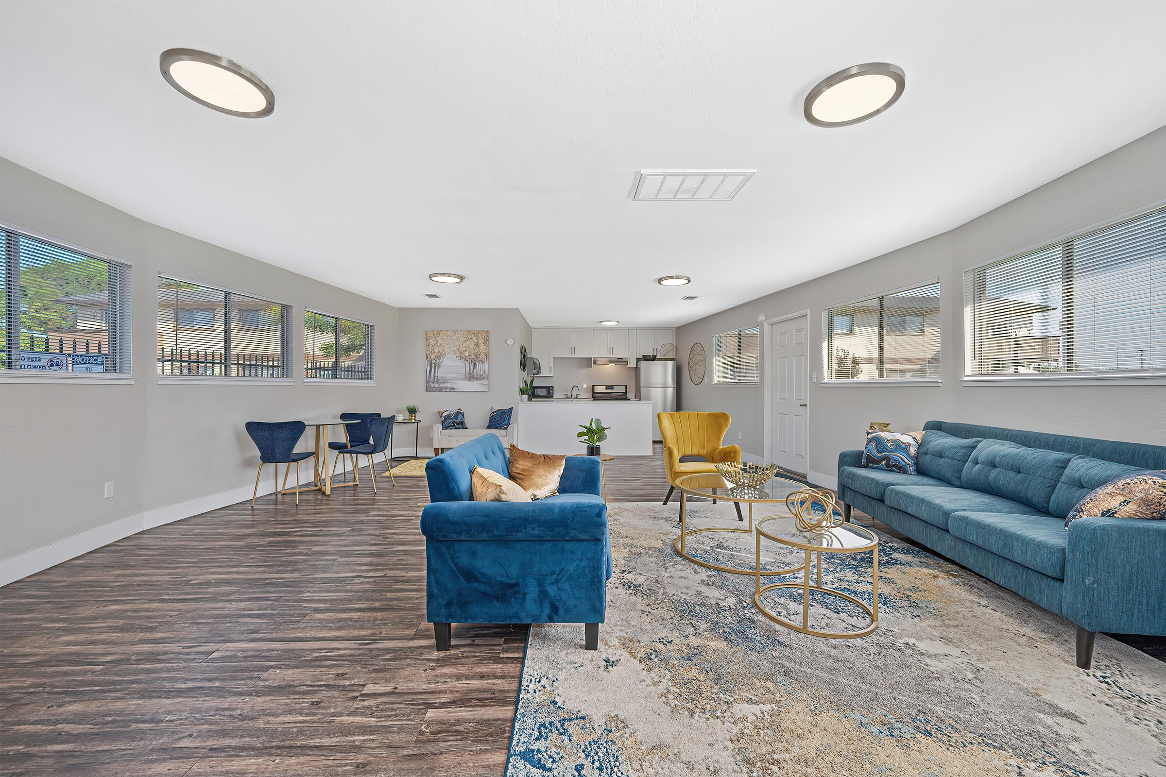 Modern living room with large windows allowing natural light. Features a blue sofa, a blue armchair, and yellow chairs. A round coffee table sits on a colorful area rug. The open layout leads to a kitchen in the background. Decorative plants and artwork enhance the ambiance.