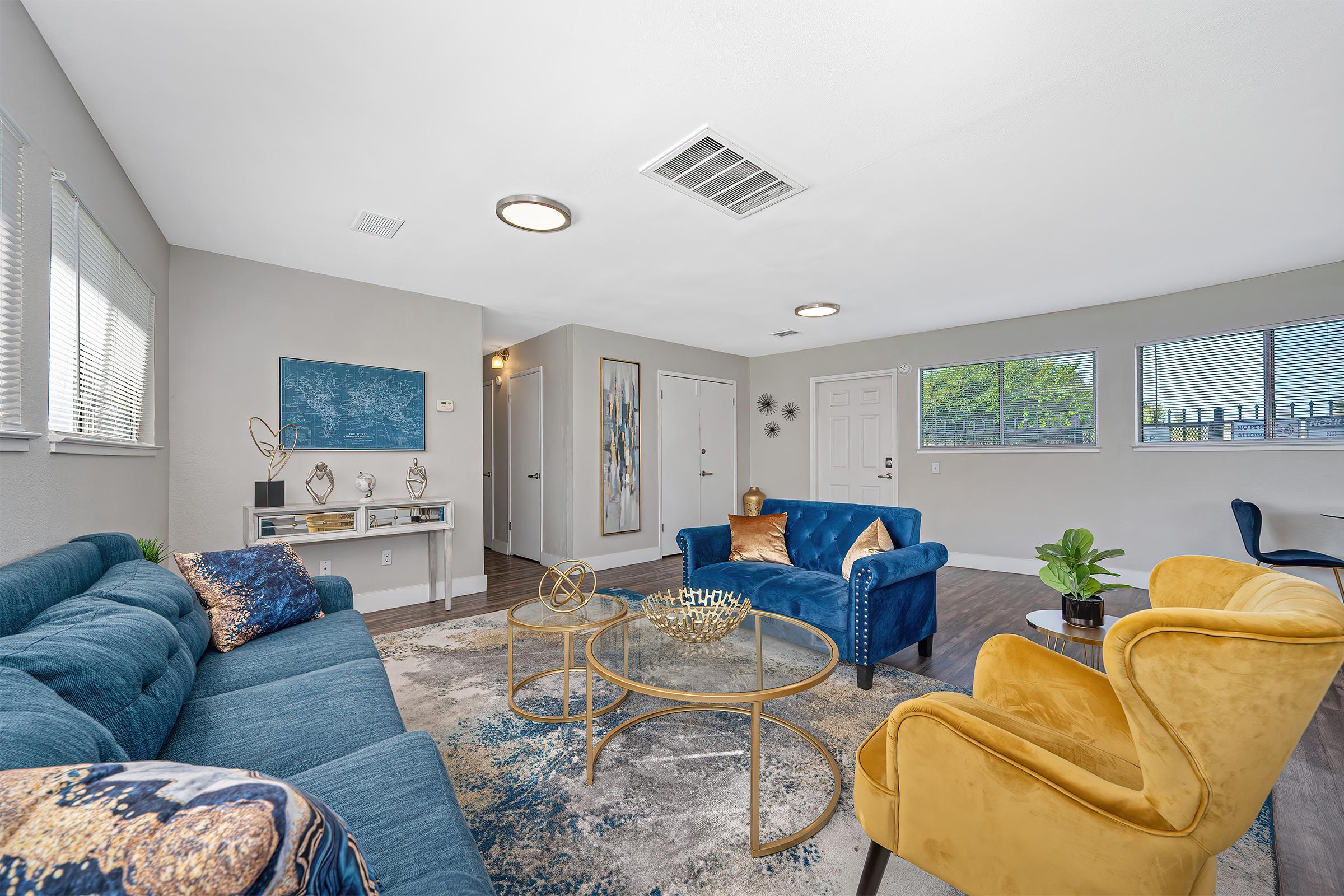 A modern living room featuring blue and yellow furniture, including a blue sofa and an orange armchair. A circular glass coffee table sits on a patterned area rug. There are large windows allowing natural light, a decorative wall art piece, and plants adding greenery to the space.