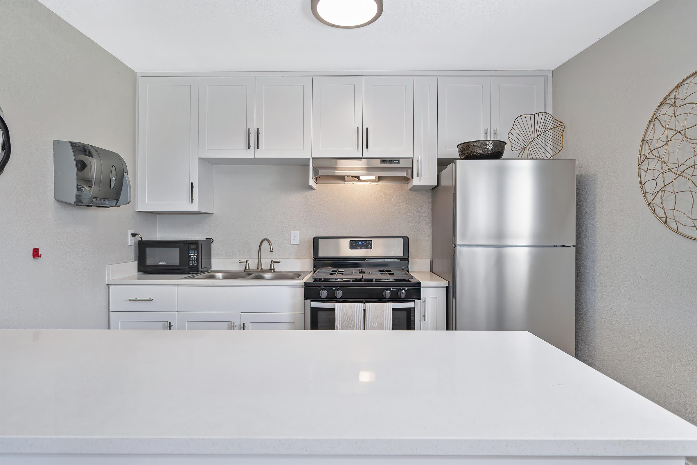 Modern kitchen featuring white cabinetry, stainless steel appliances including a refrigerator and stove, a microwave, and a sink. The countertop is sleek and white, with minimalistic decor like a round wall hanging. Soft lighting enhances the contemporary design.