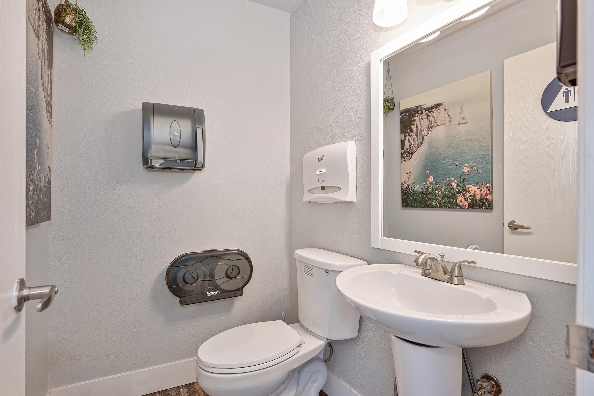 A clean, modern bathroom featuring a toilet, a sink with a faucet, and a wall-mounted paper towel dispenser. There is a mirror above the sink, and artwork depicting a coastal scene is displayed on the wall. The space is well-lit and has light gray walls.