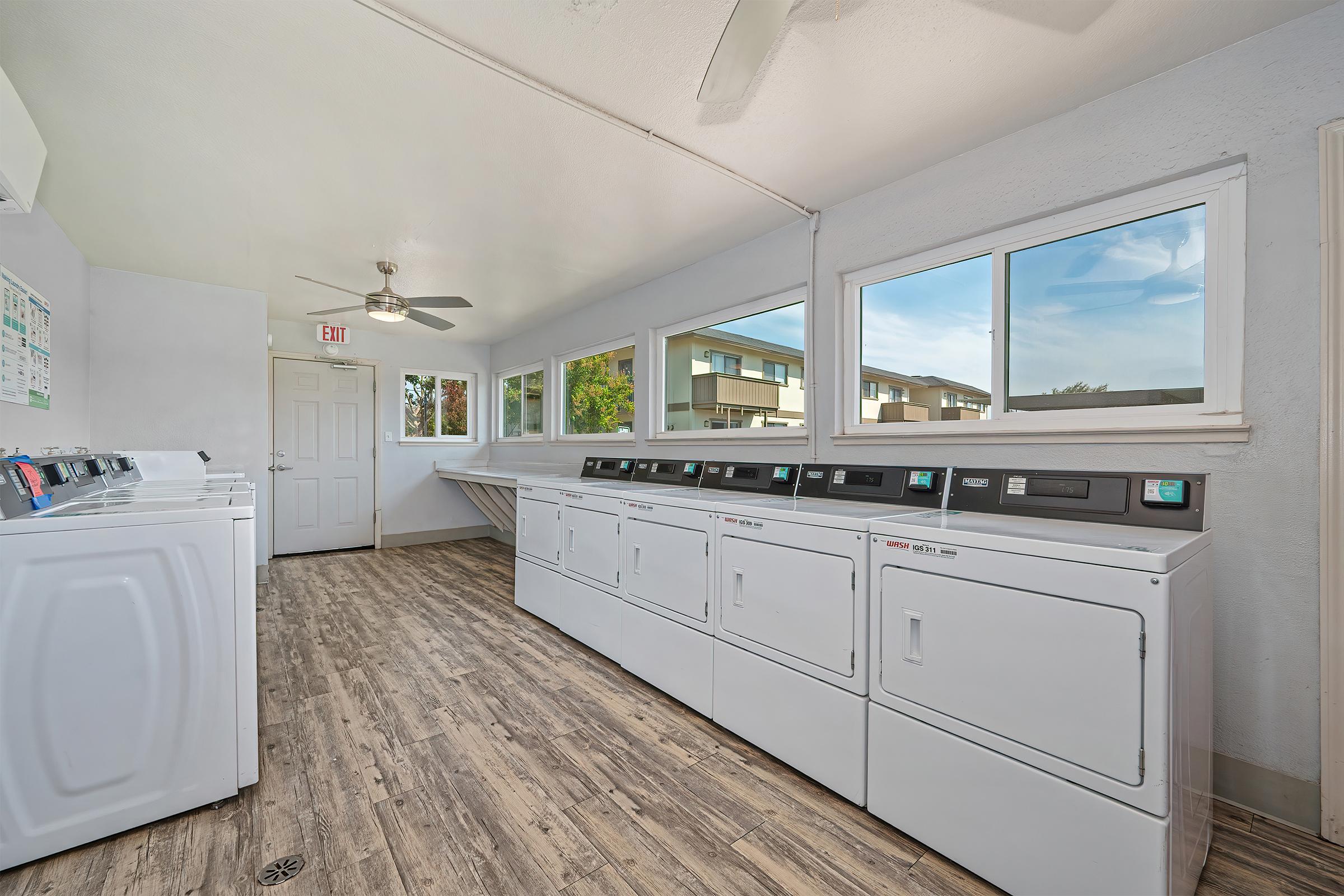 A bright and airy laundry room featuring multiple white washing machines and dryers lined up against the wall. Large windows bring in natural light, and a ceiling fan provides ventilation. A folding area is available in the corner, with an entrance door to the left. The flooring is a light wood aesthetic.