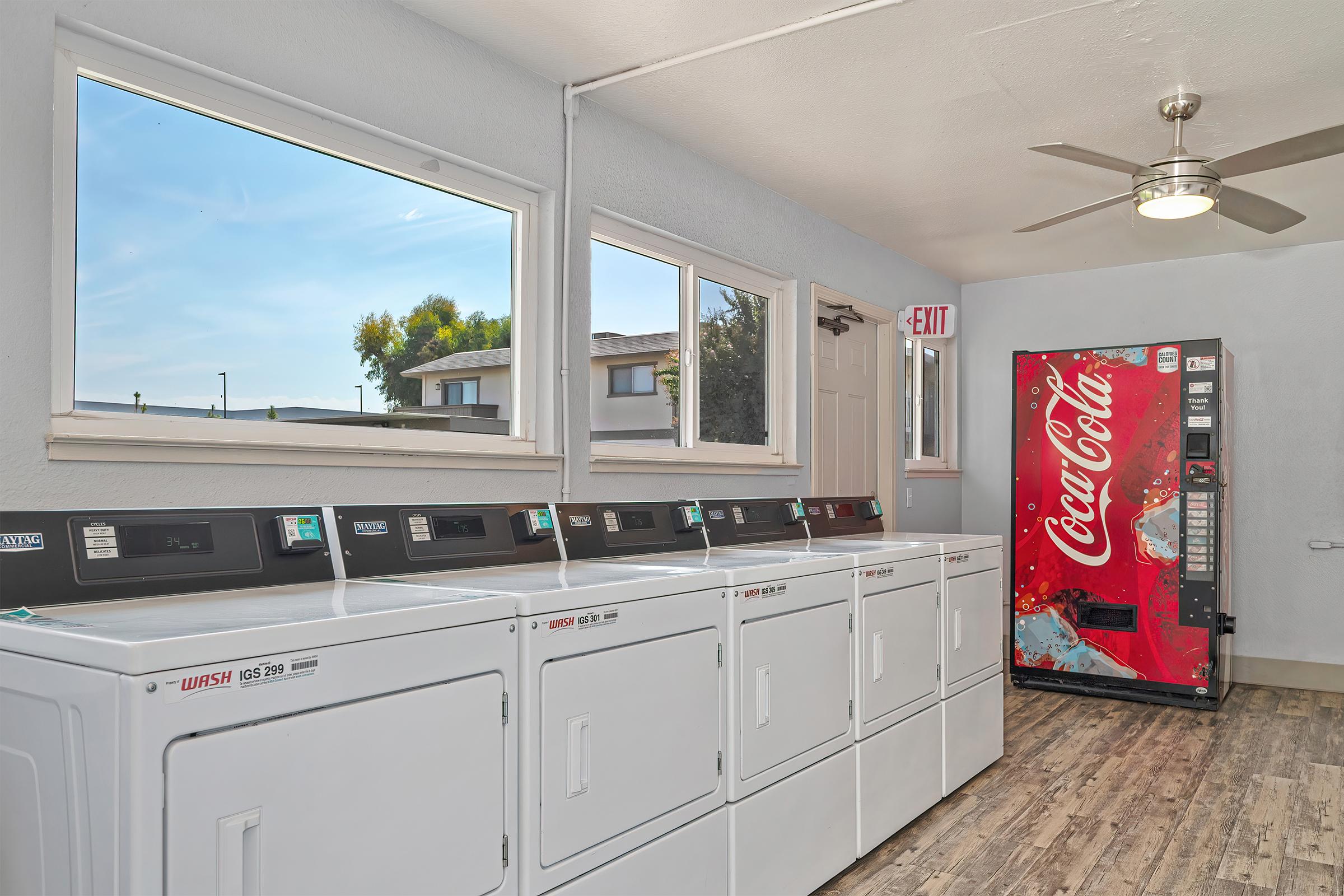 Laundry room featuring several white washing machines lined against the wall, large windows letting in natural light, a door leading outside, a ceiling fan, and a Coca-Cola vending machine in the corner. The room has a clean and modern look, with wooden flooring.