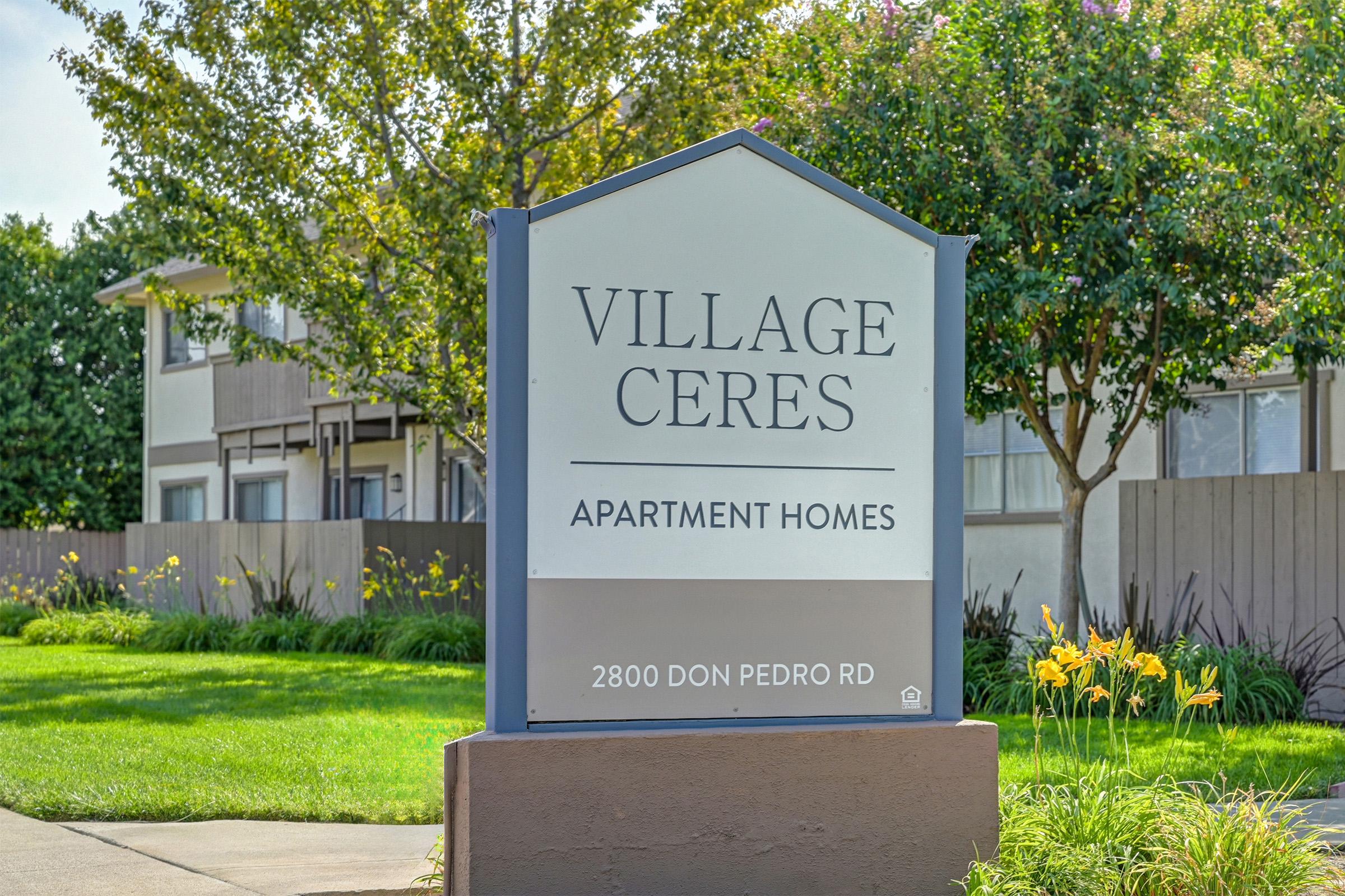 Sign for Village Ceres Apartment Homes located at 2800 Don Pedro Rd. The sign features the name "Village Ceres" prominently at the top, with "Apartment Homes" below it. Surrounding landscaping includes greenery and flowers, providing a welcoming atmosphere.