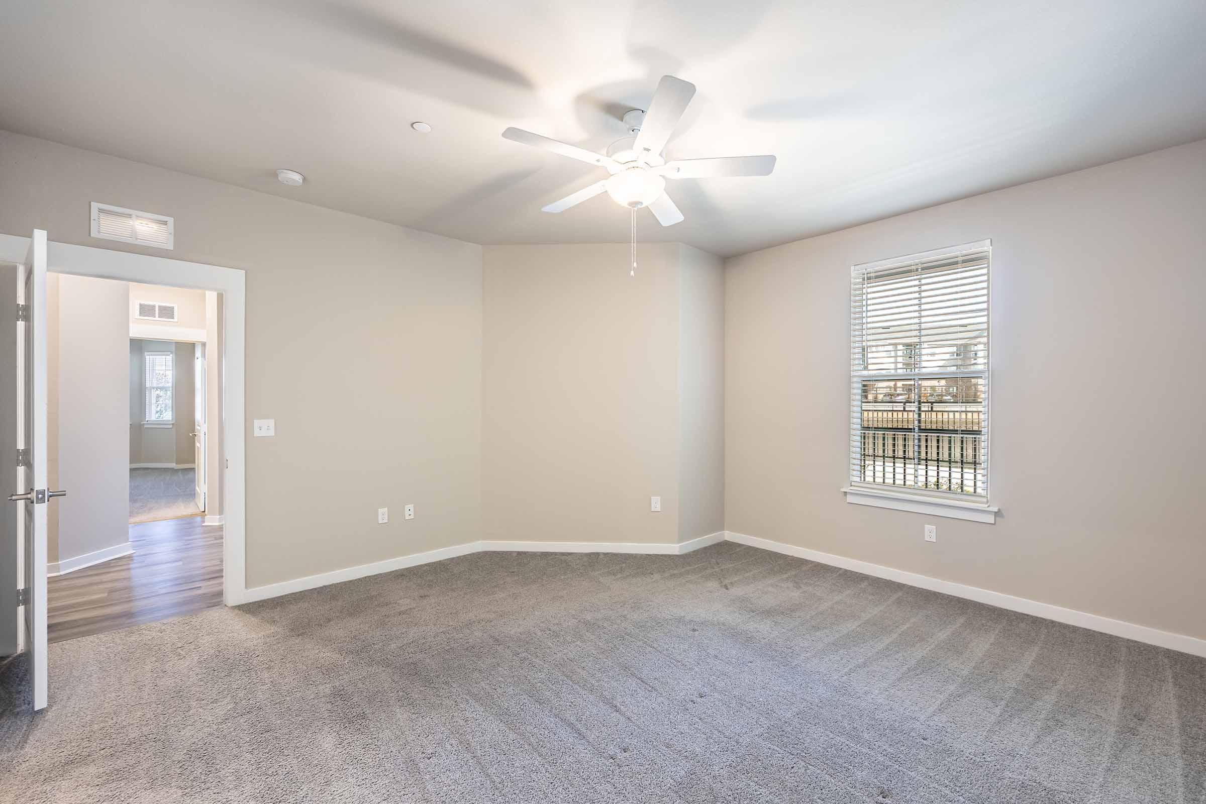 A spacious, empty room with light-colored walls and carpet. A ceiling fan is mounted above, and there is a window with blinds allowing natural light to enter. A door leads to another room in the background, creating a bright and airy atmosphere.