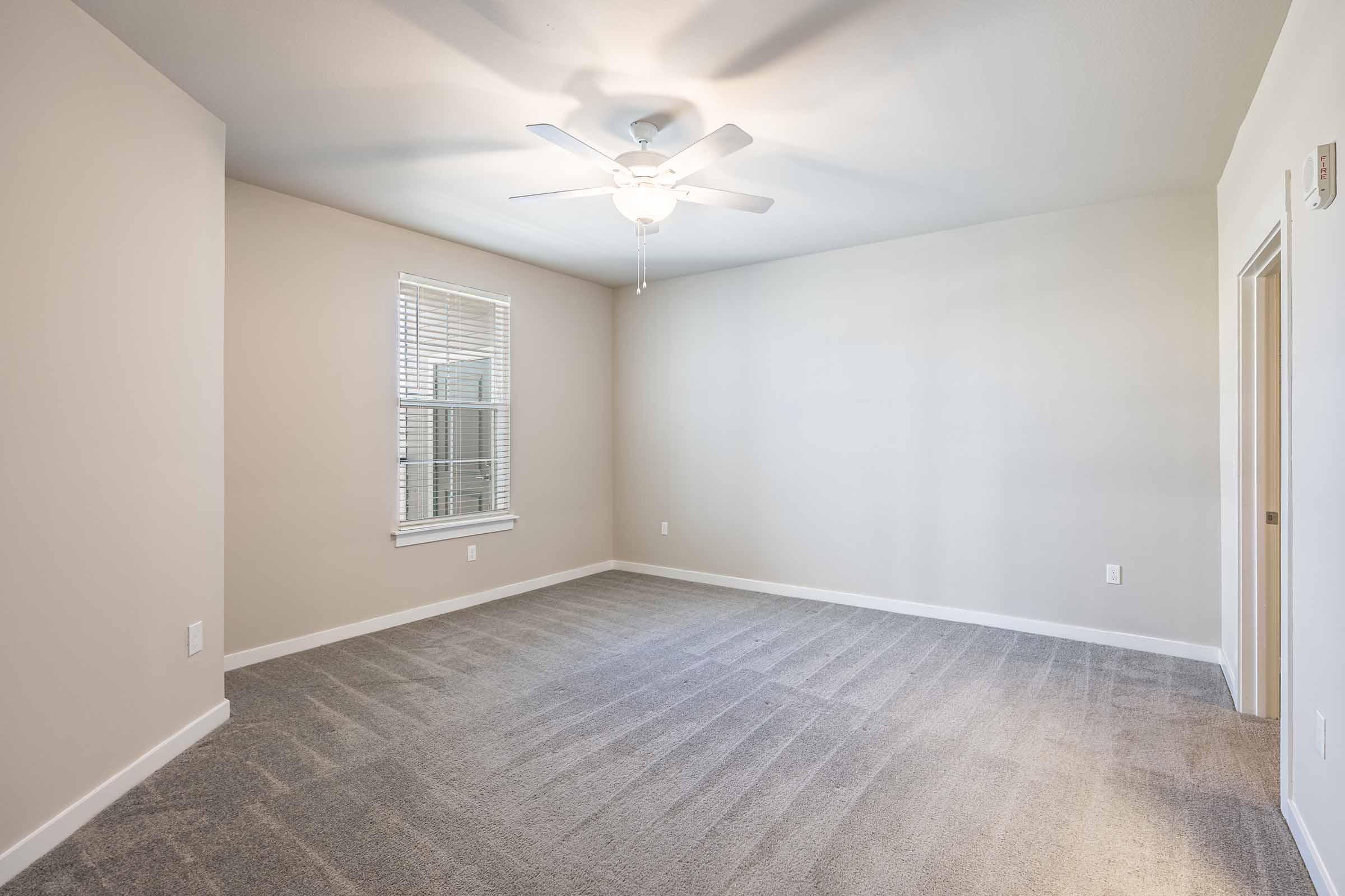 A spacious, empty room with light-colored walls and a ceiling fan. The floor is covered in plush carpeting. Large windows allow natural light to enter, highlighting the room’s openness. No furniture is present, creating a clean, uncluttered look.