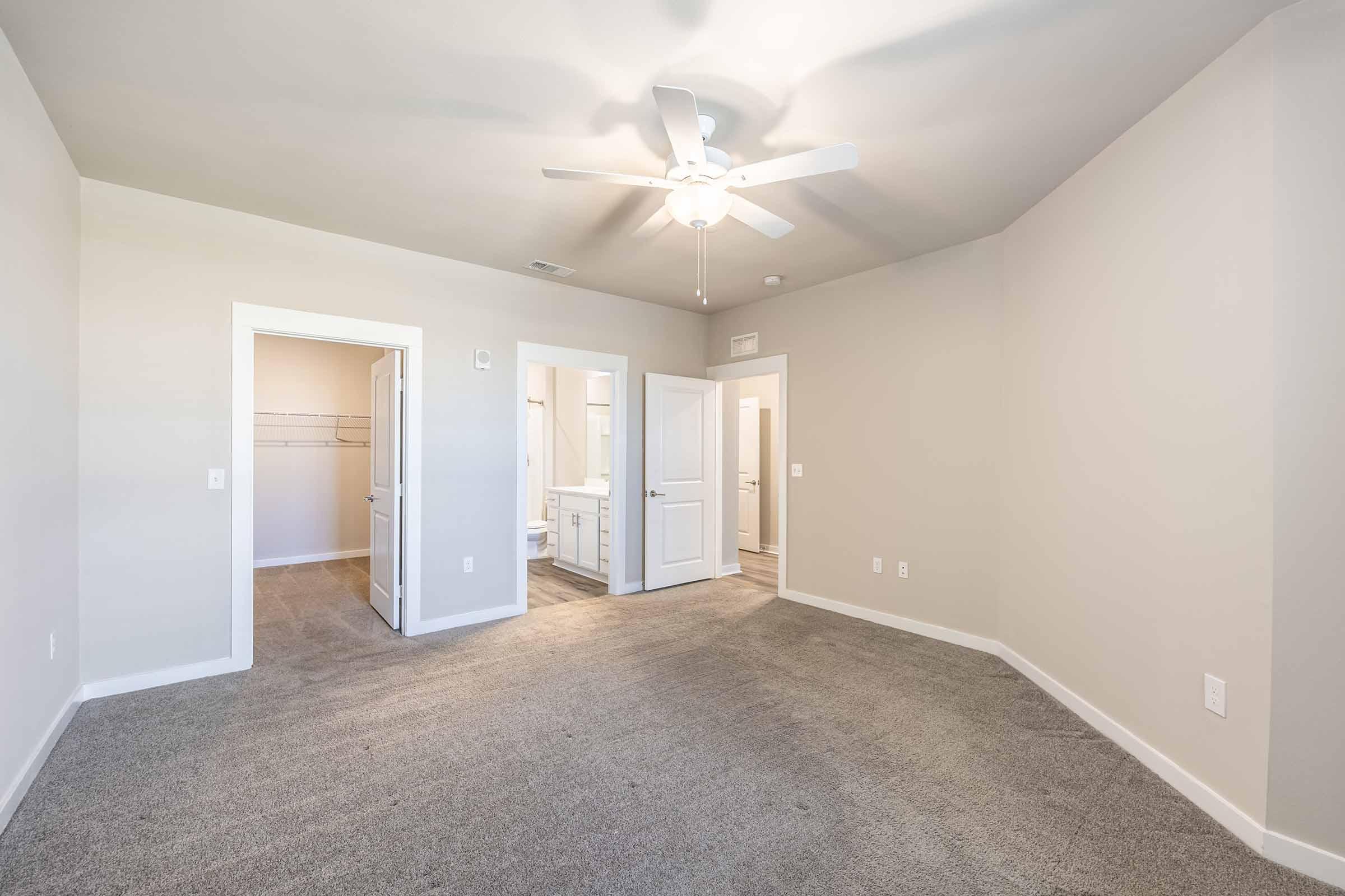 A spacious room with light gray walls and carpeted flooring. It features a ceiling fan and opens to two doorways; one leading to a bathroom and the other to a hallway. Natural light illuminates the space, creating a bright and airy atmosphere. The room is unoccupied and has no visible furniture.