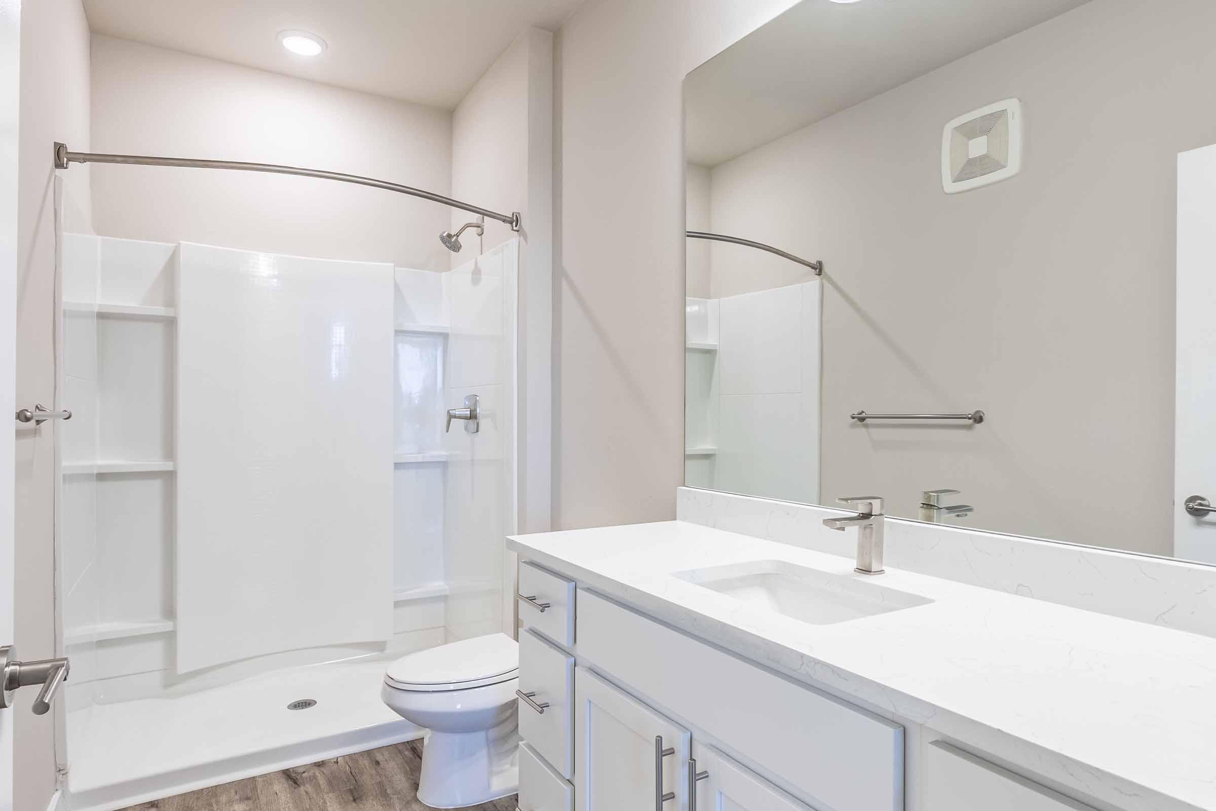 A modern bathroom featuring a white shower with a curved curtain rod, a toilet, and a double-sink vanity with a sleek countertop. The walls are painted in a light neutral color, and there are bright overhead lights. A large mirror above the vanity complements the clean and contemporary design.
