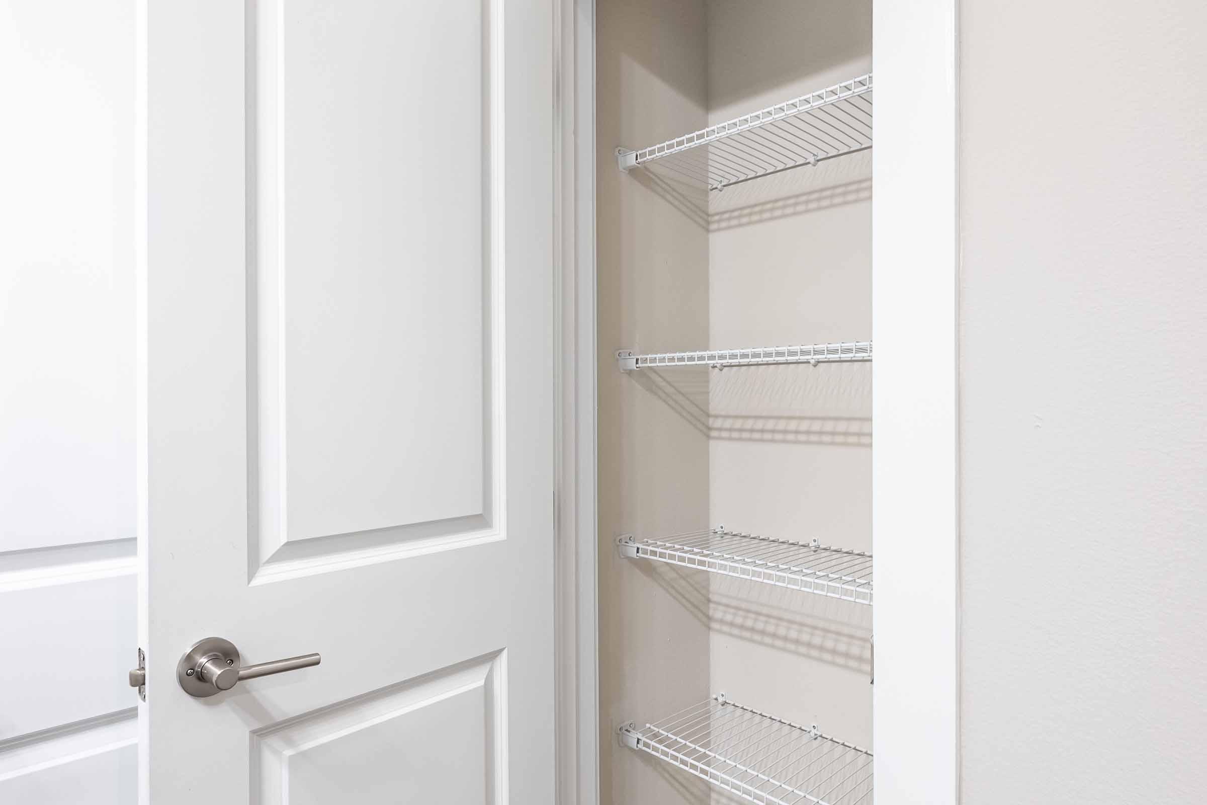 A white door partially open, revealing a closet with wire shelving. The closet has four shelves and a light-colored wall with a clean, minimalist design. The door handle is silver, and the overall aesthetic is modern and tidy.