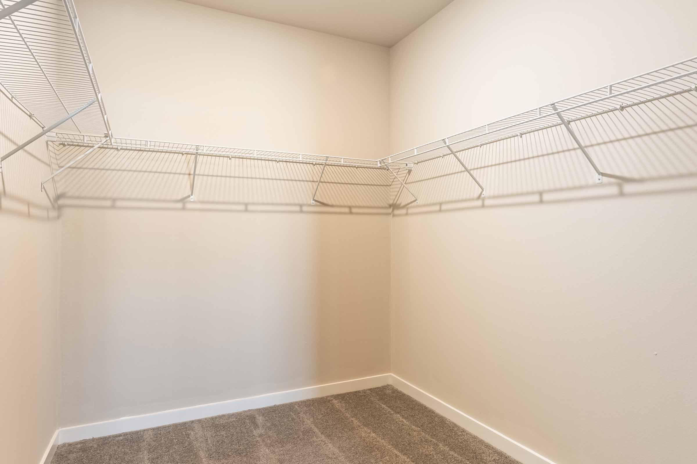 Empty closet with white wire shelving mounted on the walls. The walls are painted a light color, and the floor is covered with a soft, neutral-toned carpet. The overall space appears clean and minimally furnished, providing ample room for storage.