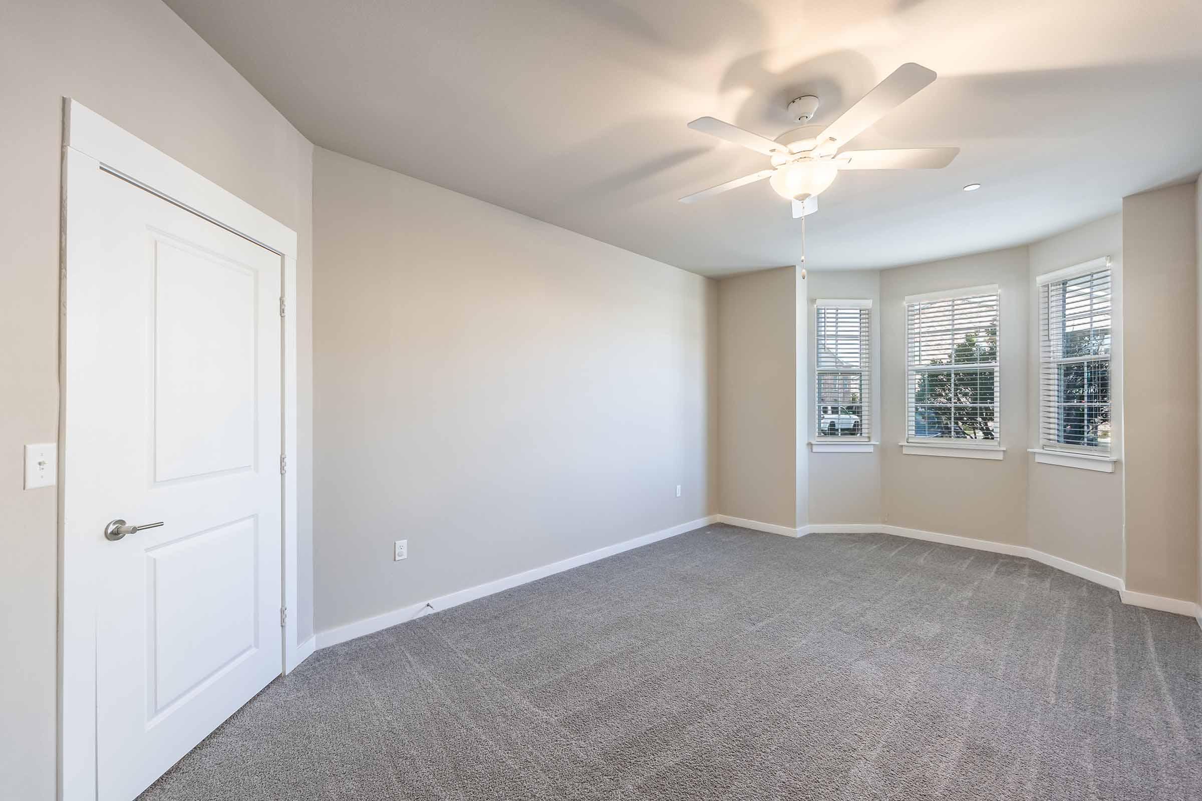 A spacious, empty room featuring light gray walls and carpet. It has a white ceiling fan, a large window with multiple panes allowing natural light, and a white door. The room is neatly designed with a modern feel, making it suitable for various purposes like a bedroom or office.