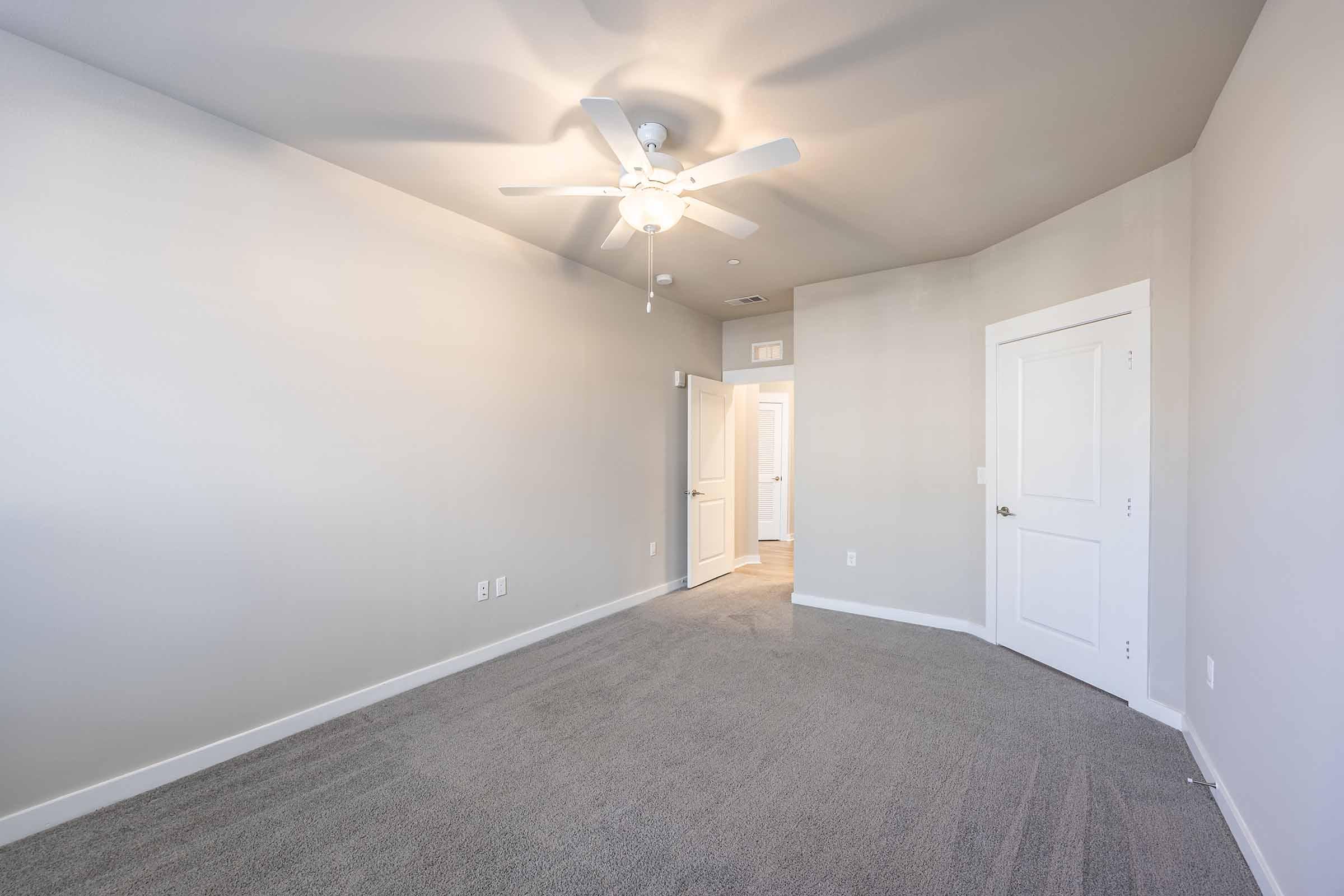 A spacious empty room featuring light gray walls, a ceiling fan, and soft gray carpet. The room has a doorway leading to another area, with bright light coming from an adjacent space, creating a welcoming atmosphere.