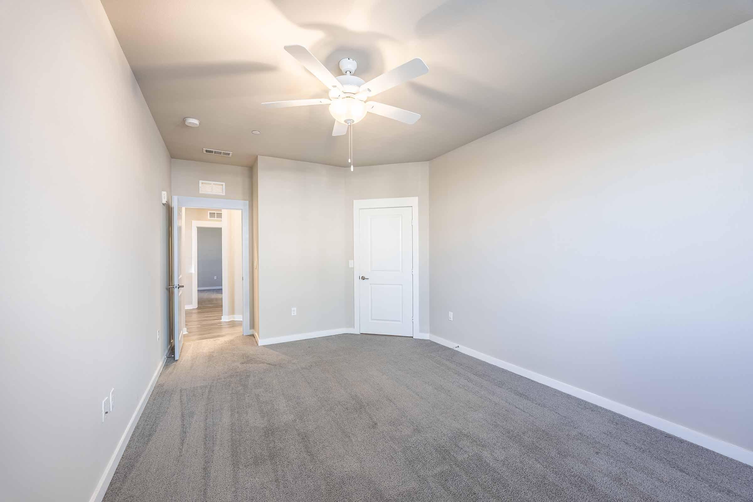 Empty room with light-colored walls, a ceiling fan, and plush grey carpet. There is a doorway leading to another space, and natural light fills the room, creating a bright and inviting atmosphere. The layout is simple and clean, suitable for various interior design possibilities.