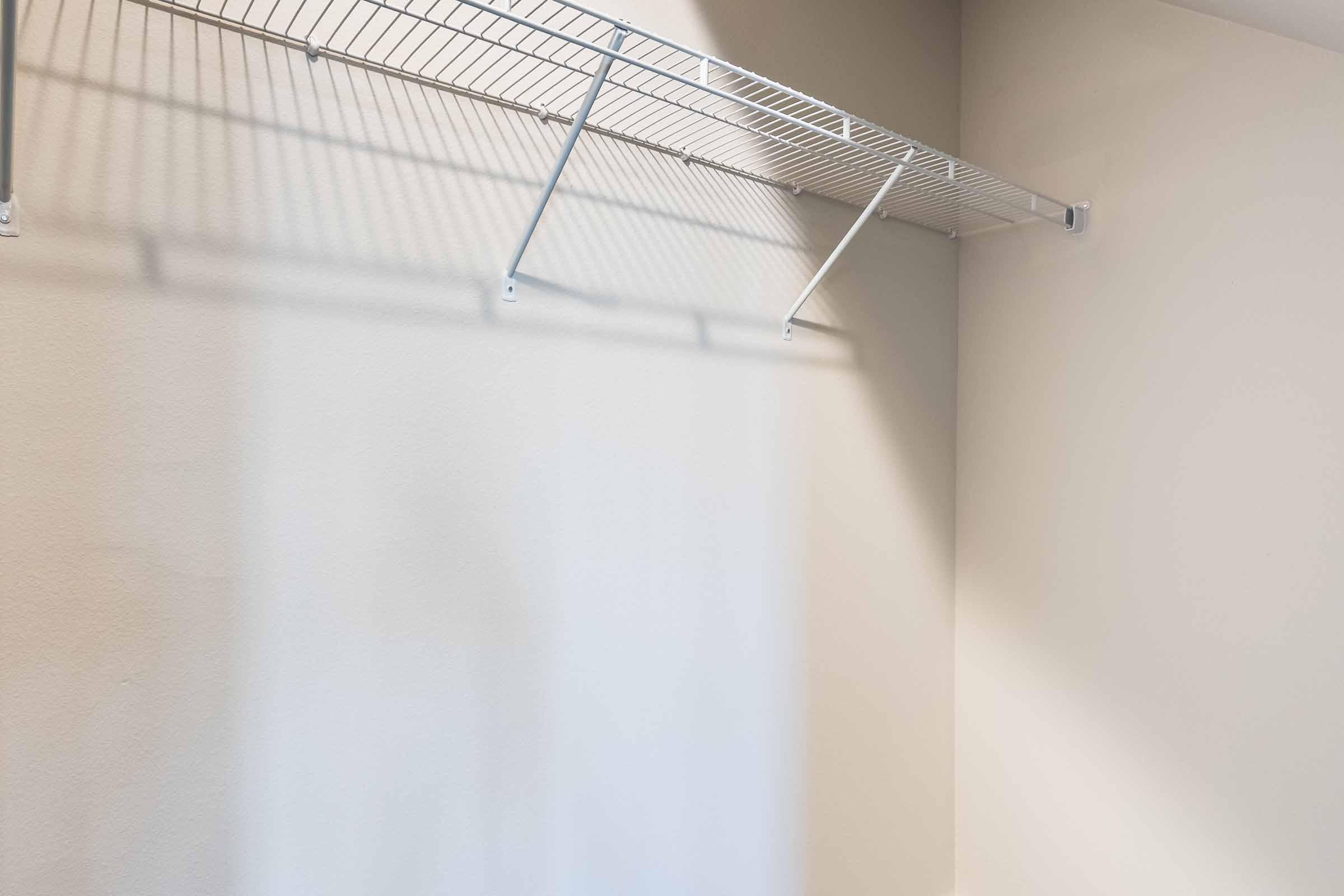 A blank wall with a wire shelving unit mounted near the top. The shelf is empty and casts shadows on the wall, suggesting a minimalist or unused storage space. The surrounding area is well-lit and has a neutral color scheme.