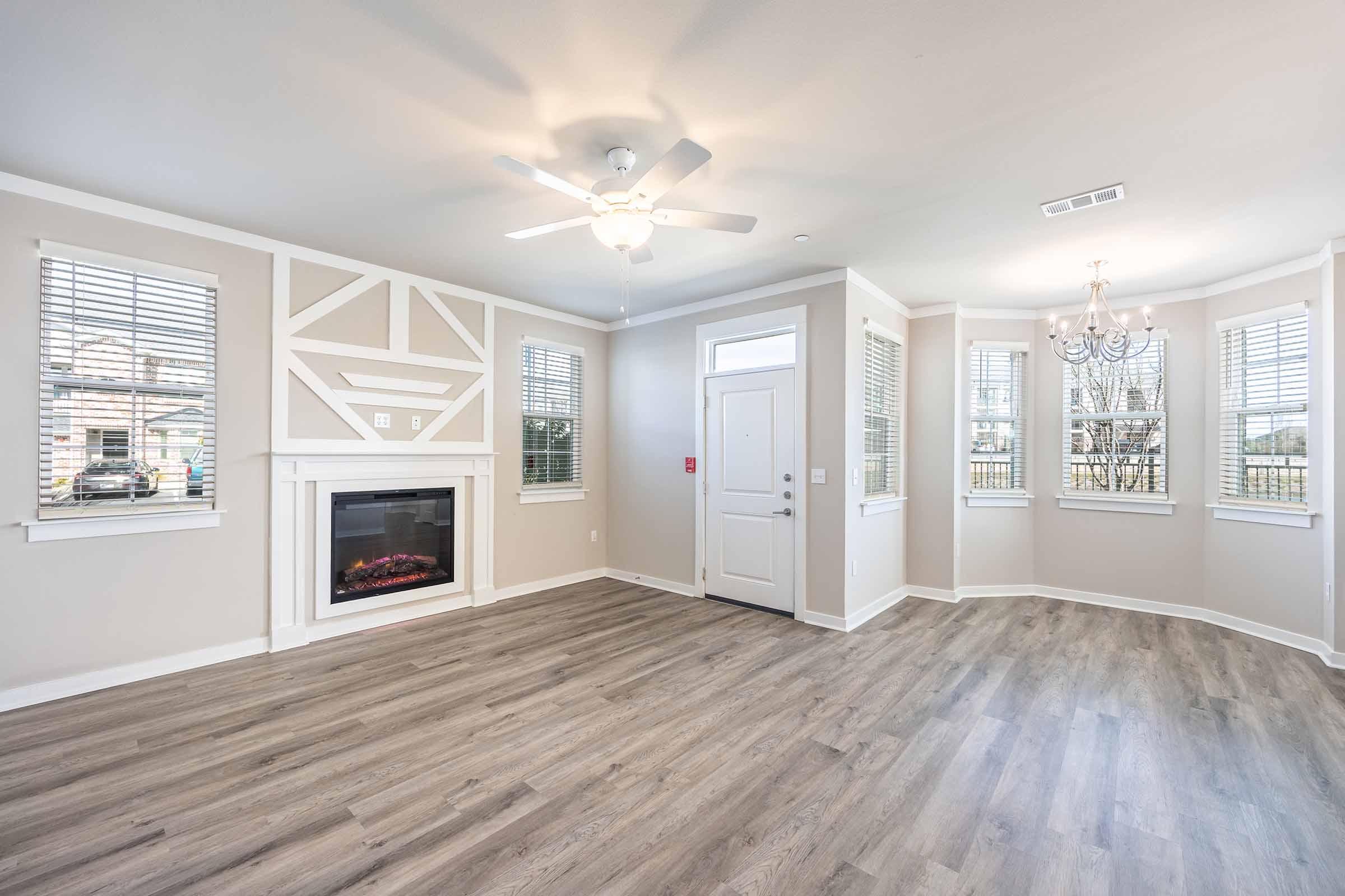 Bright and spacious living room with light-colored walls and elegant trim. Features a modern ceiling fan, a cozy electric fireplace, large windows with blinds, and minimalist decor. The room is illuminated by natural light, showcasing a stylish chandelier and warm wooden flooring.