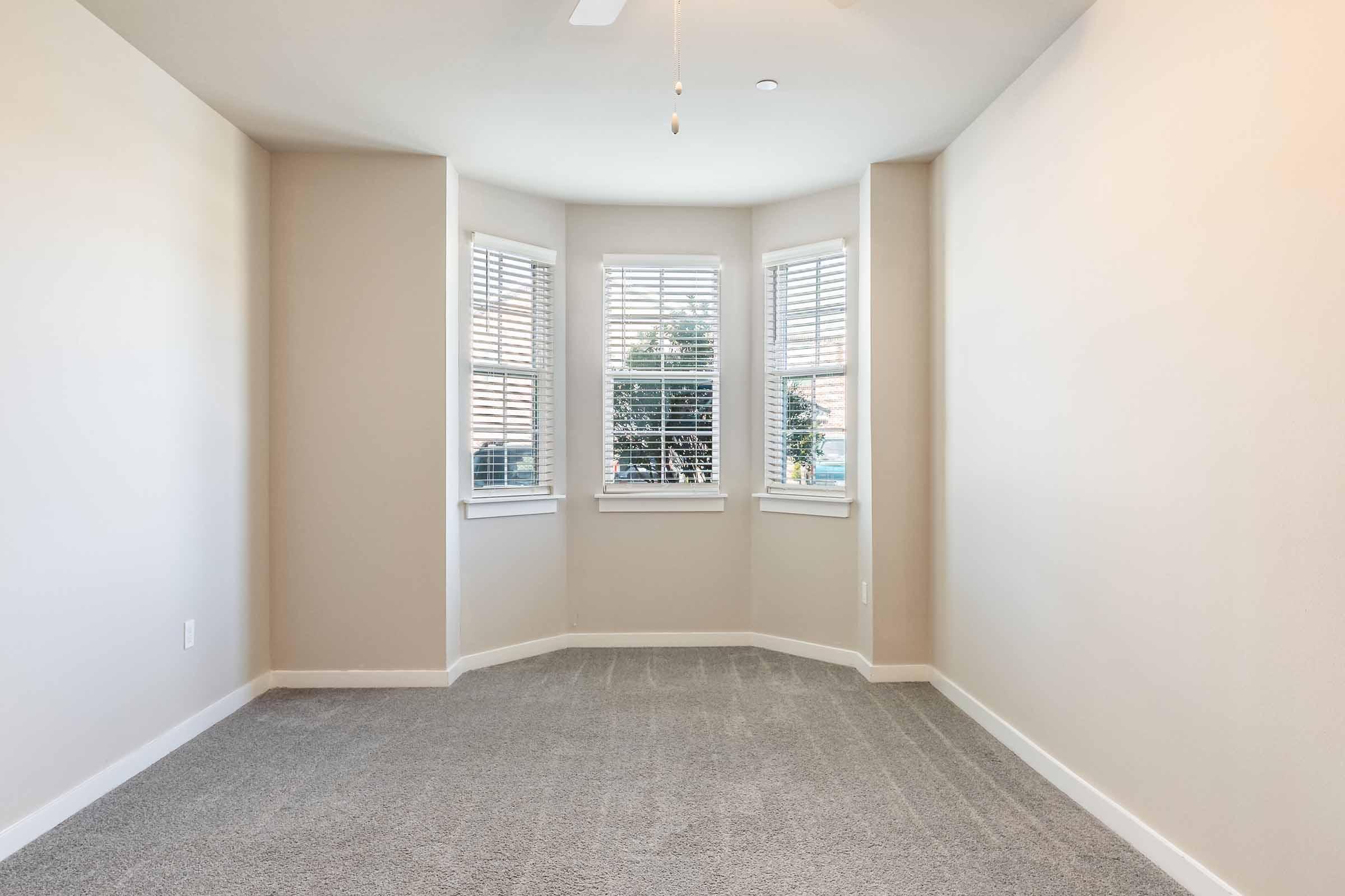 A spacious empty room with light gray carpet and a neutral-colored wall. Large windows with white blinds provide natural light, and a ceiling fan is visible. The room is designed with a bay window feature, offering a cozy nook for seating or decor.