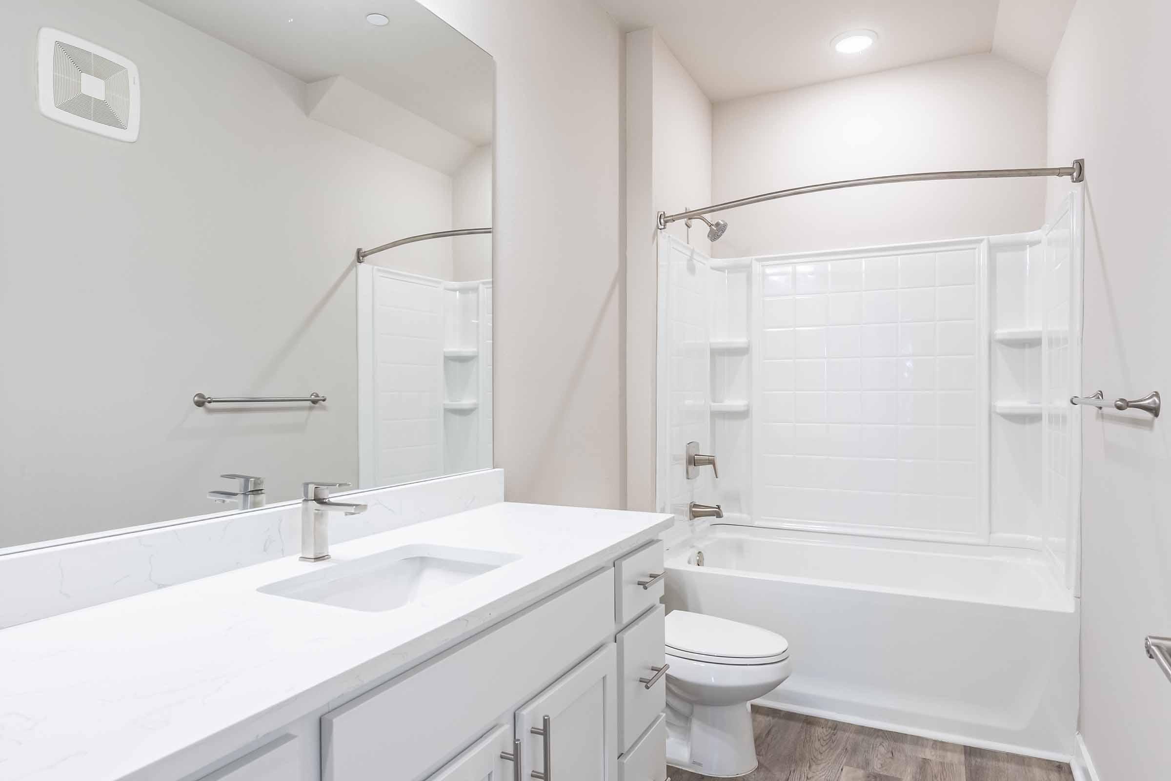 A modern bathroom featuring a white bathtub with a shower, a sleek white sink with a brushed nickel faucet, and a large mirror above the sink. The walls are painted in a light color, and the floor has a wood-like texture. Soft lighting enhances the bright, clean appearance of the space.